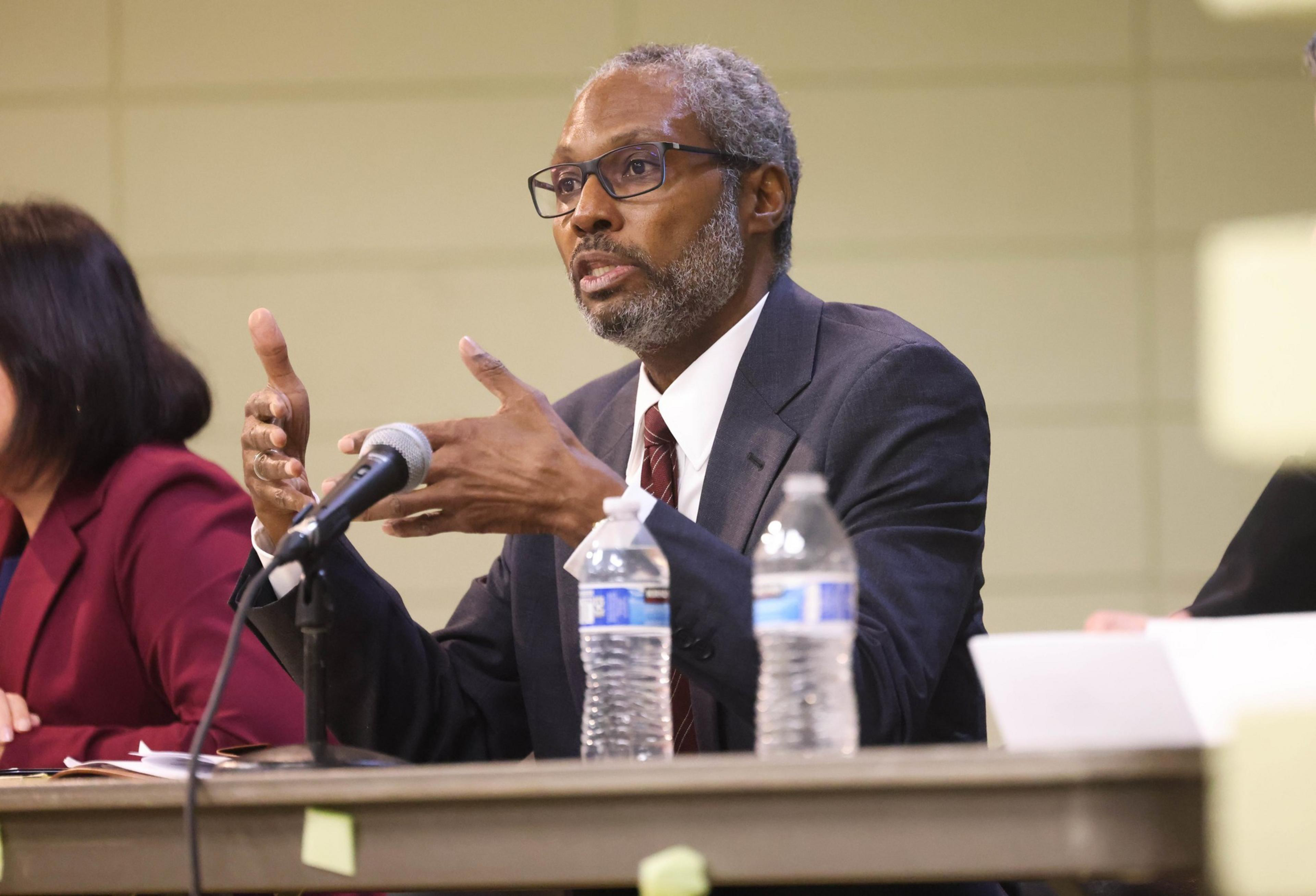 A man sits at a table talking to an audience. 