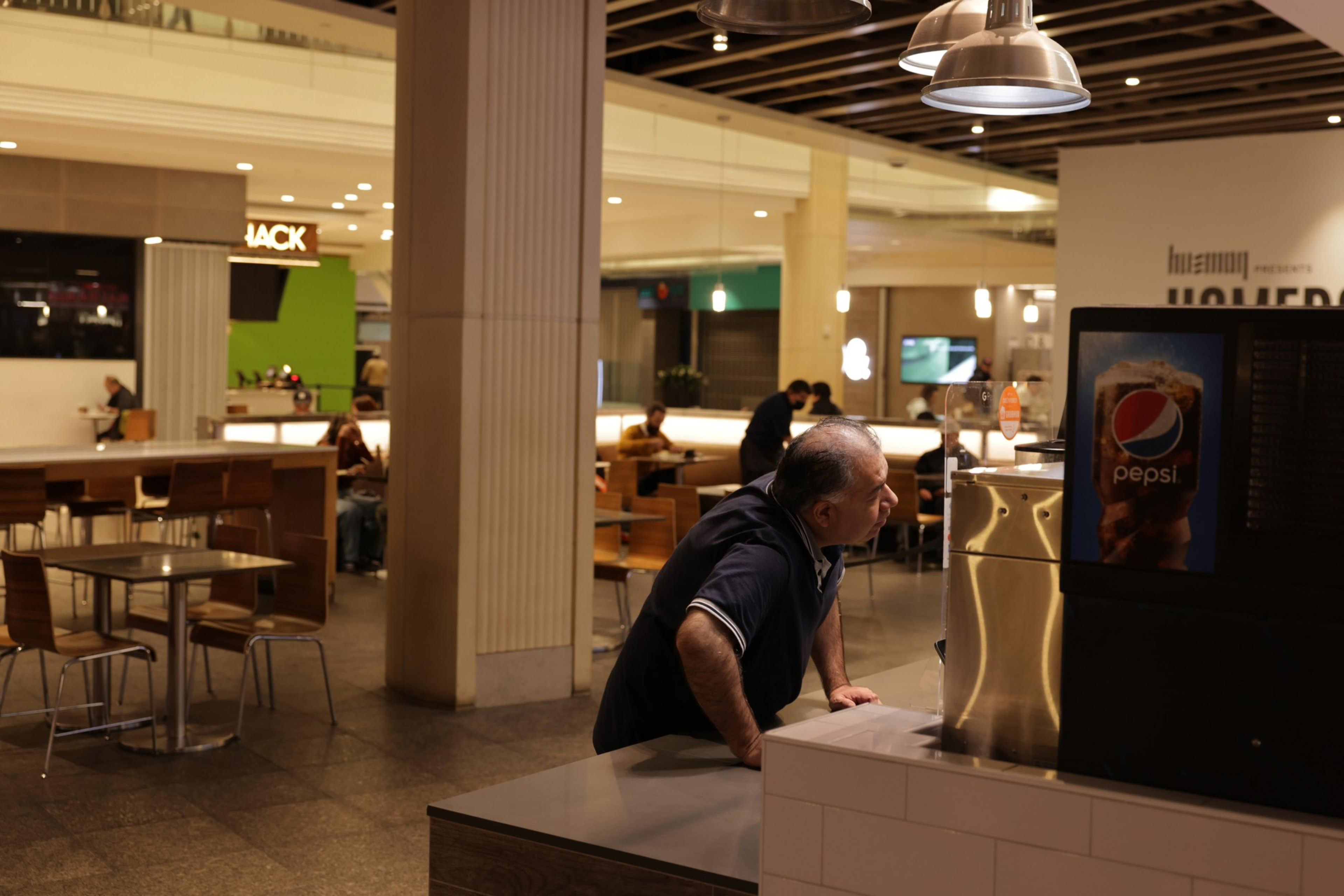 A man leans on a counter in a dimly lit food court, looking away thoughtfully. Tables, diners, and a Pepsi ad are visible.