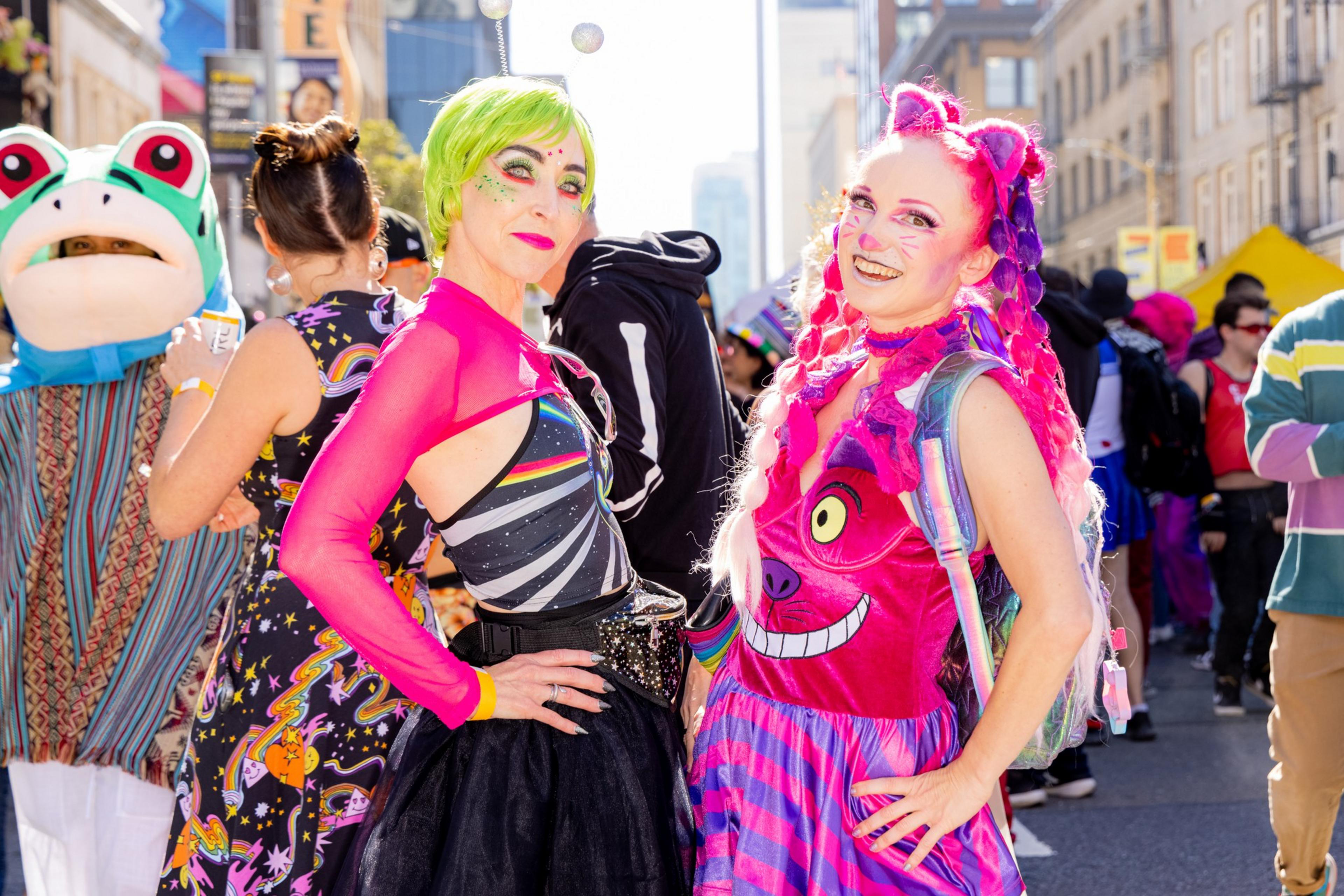 Two people in vibrant costumes pose; one with green hair and sparkly makeup, the other with pink hair and a Cheshire Cat dress. A person in a frog costume and other colorful characters are in the background.