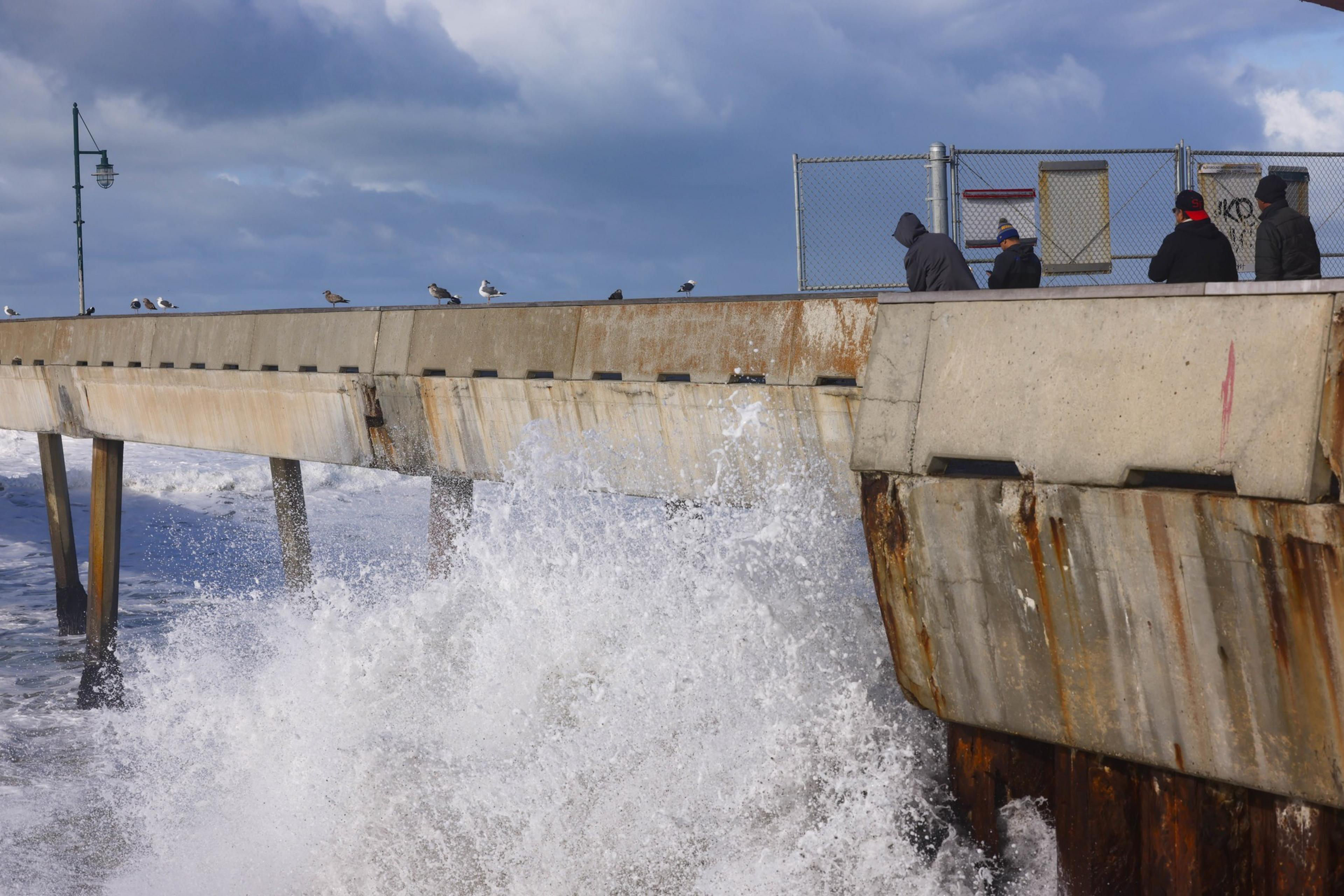 People look on as waves crash against a concrete pier