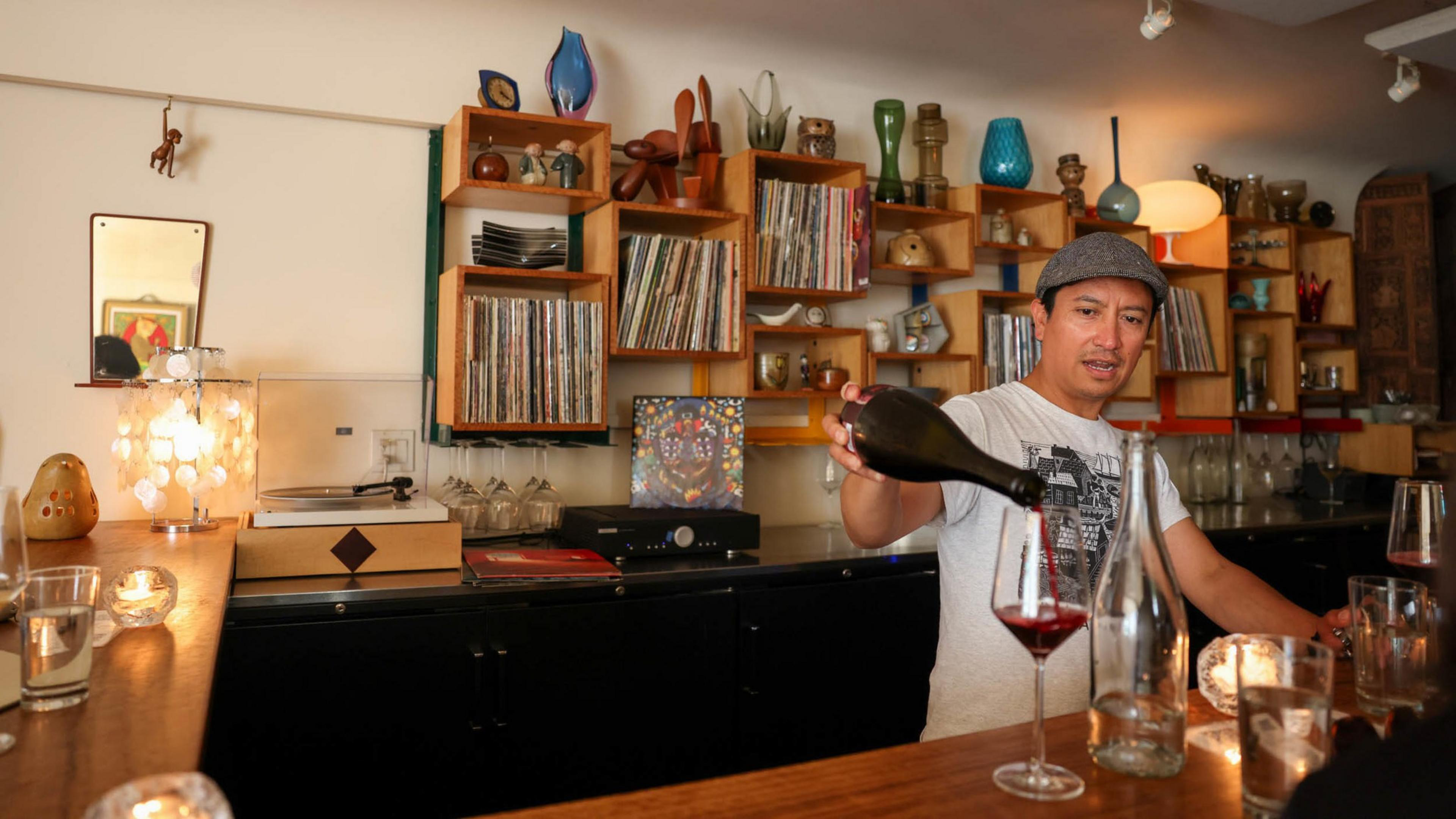 A man wearing a gray hat pours red wine into a glass behind a wooden bar lined with candles, glassware, and a turntable with vinyl records on shelves.