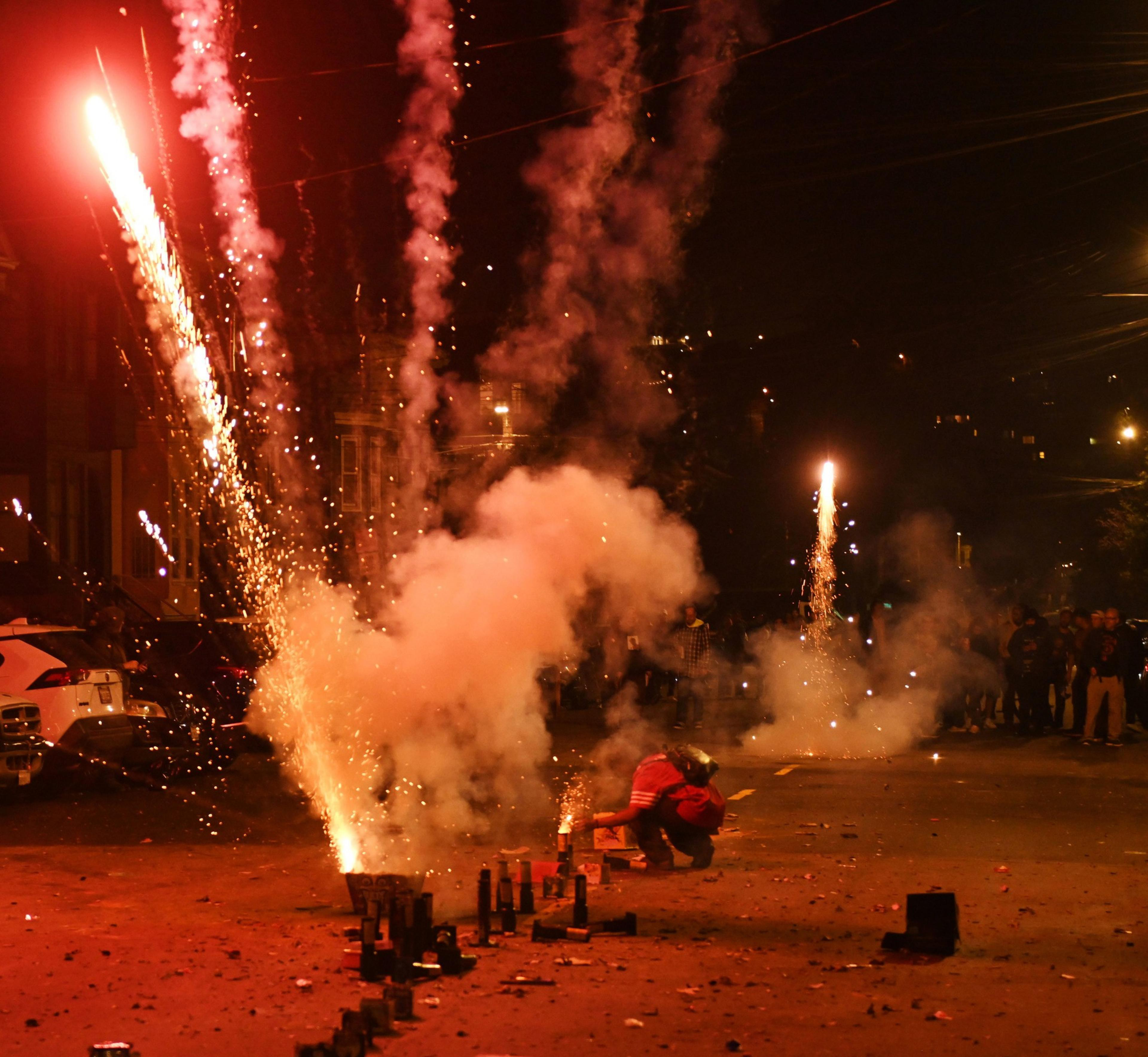 A night scene with fireworks shooting and illuminating the sky; a person kneels close to the sparks, surrounded by smoke, while onlookers watch from a distance.