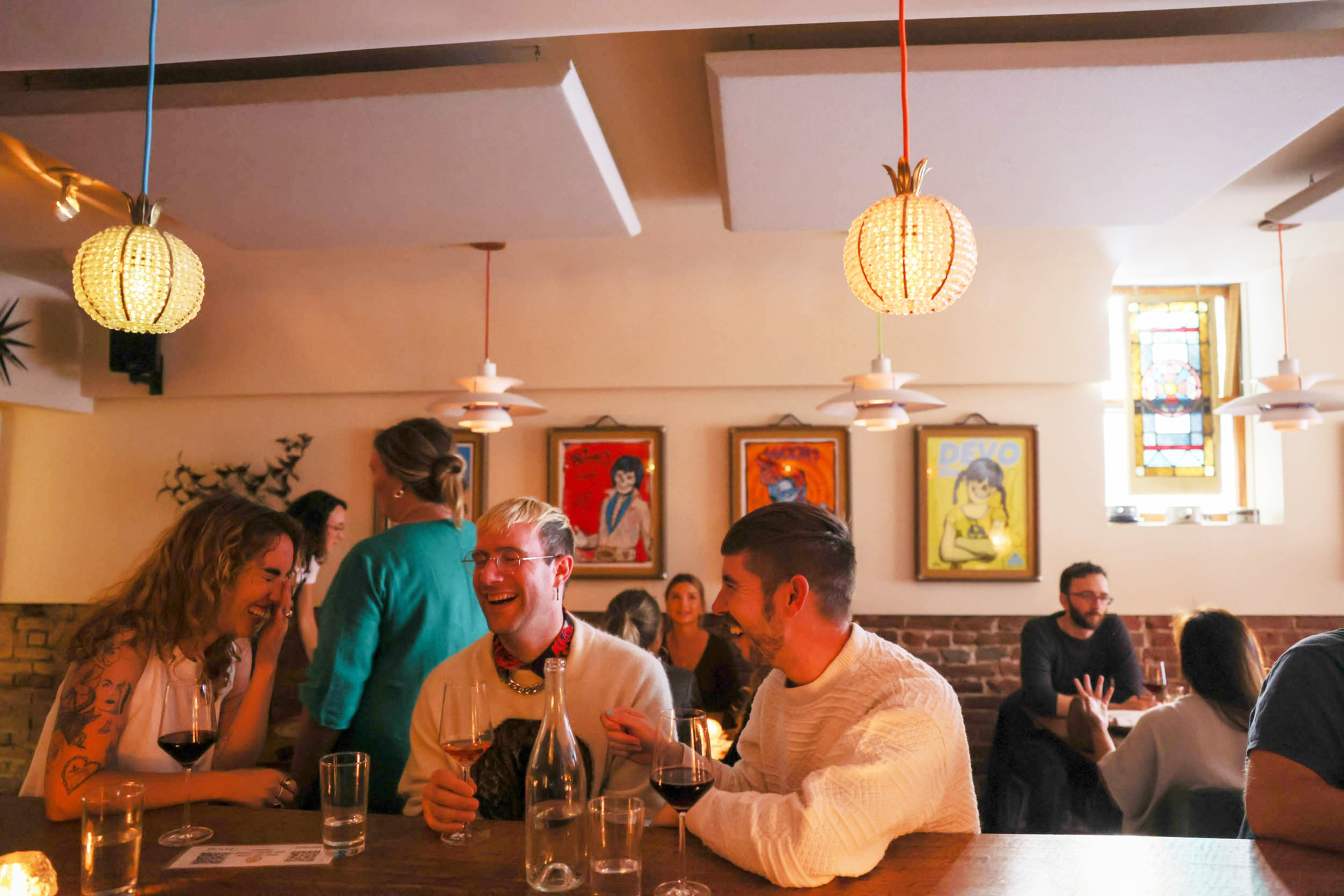 People are laughing and chatting at a cozy bar with warm lighting, decorative lamps, and colorful artwork on the walls. Wine glasses are on the table.