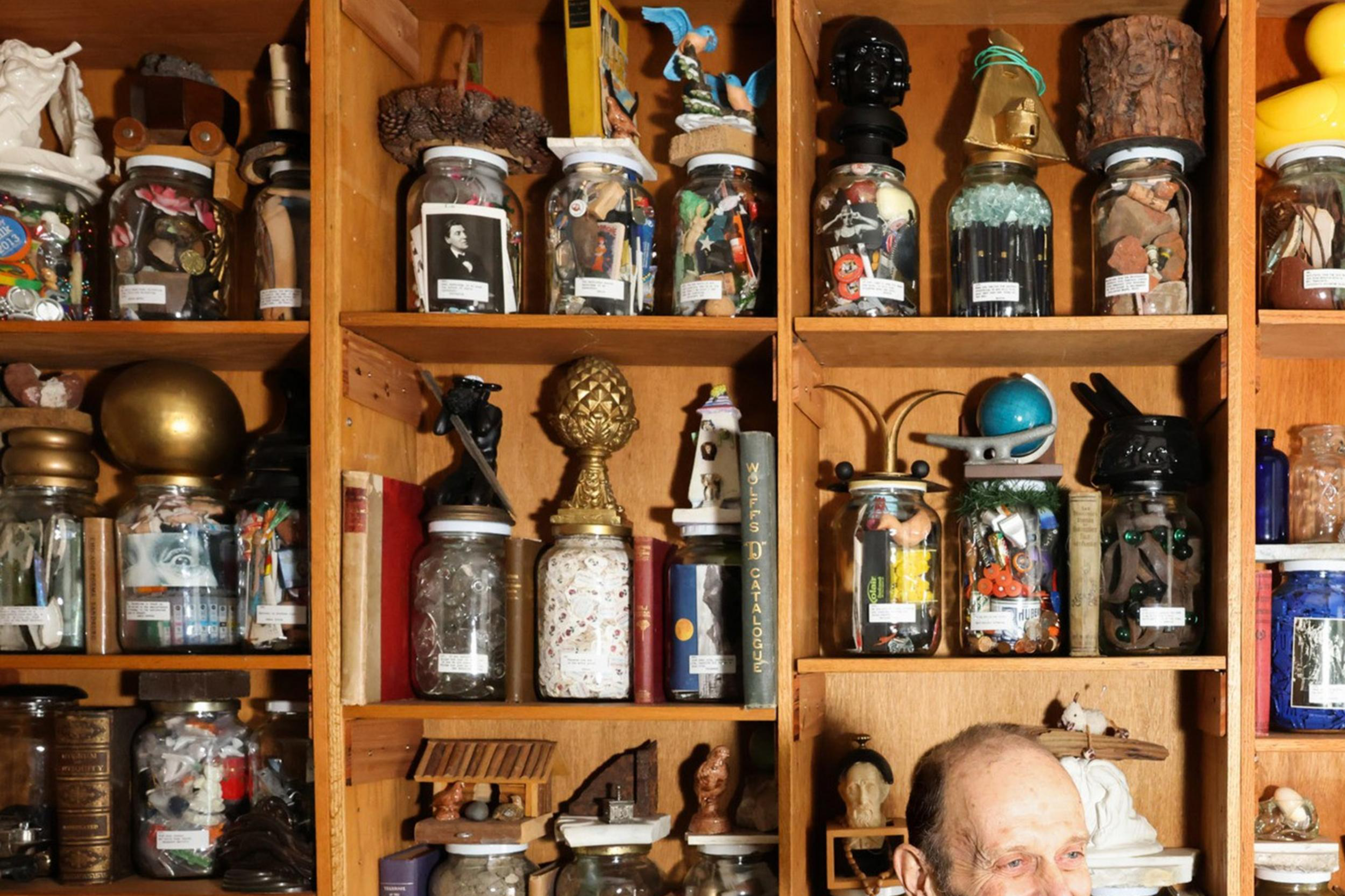 A man smiles in front of wooden shelves filled with various jars, each containing eclectic items like toys, artifacts, and decorative objects.
