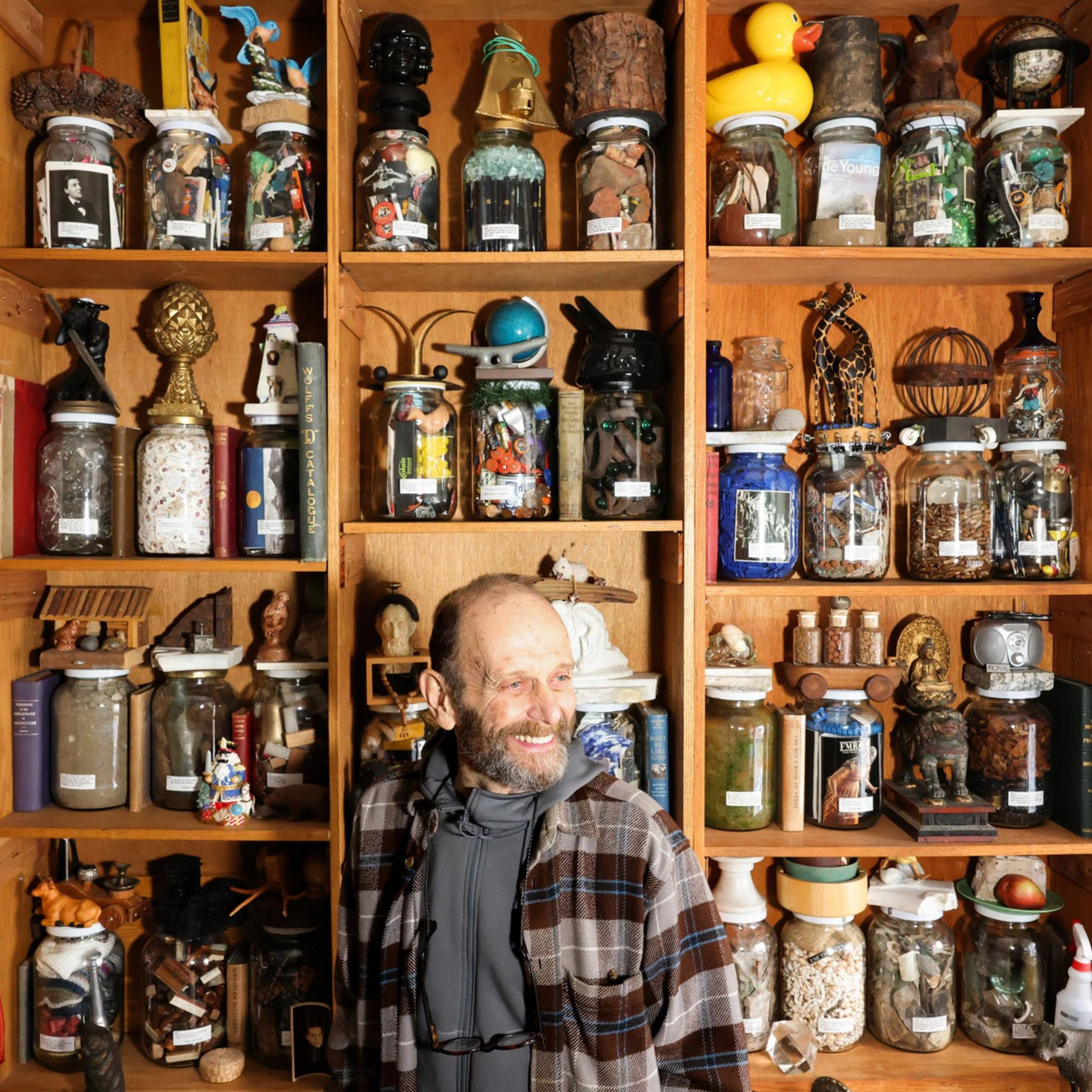 A man smiles in front of wooden shelves filled with various jars, each containing eclectic items like toys, artifacts, and decorative objects.