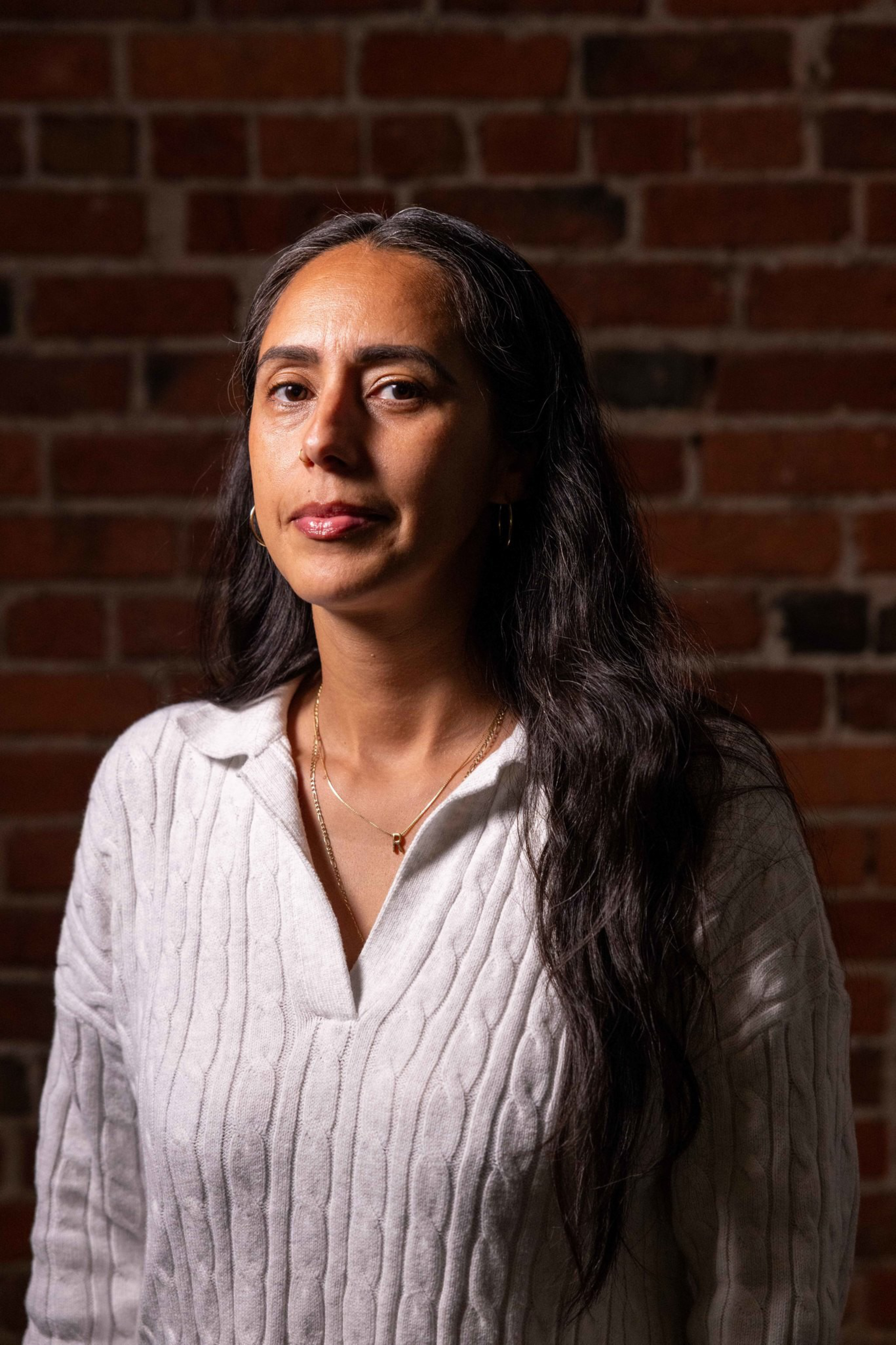 A woman with long, dark hair wears a white cable-knit sweater and gold jewelry, standing against a brick wall with a neutral expression.
