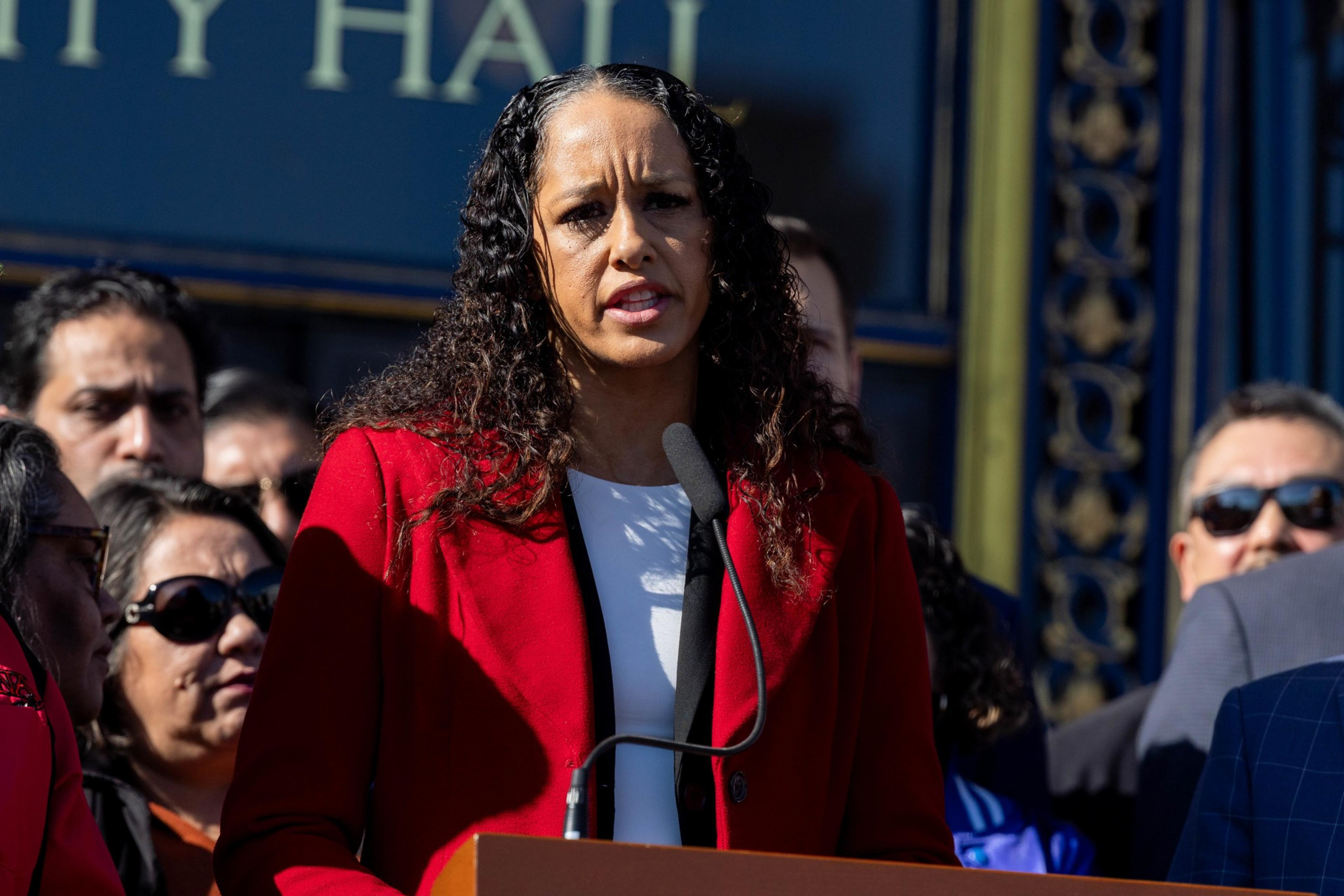 A woman in a red jacket speaks at a microphone in front of a crowd. The background features a building with intricate details.