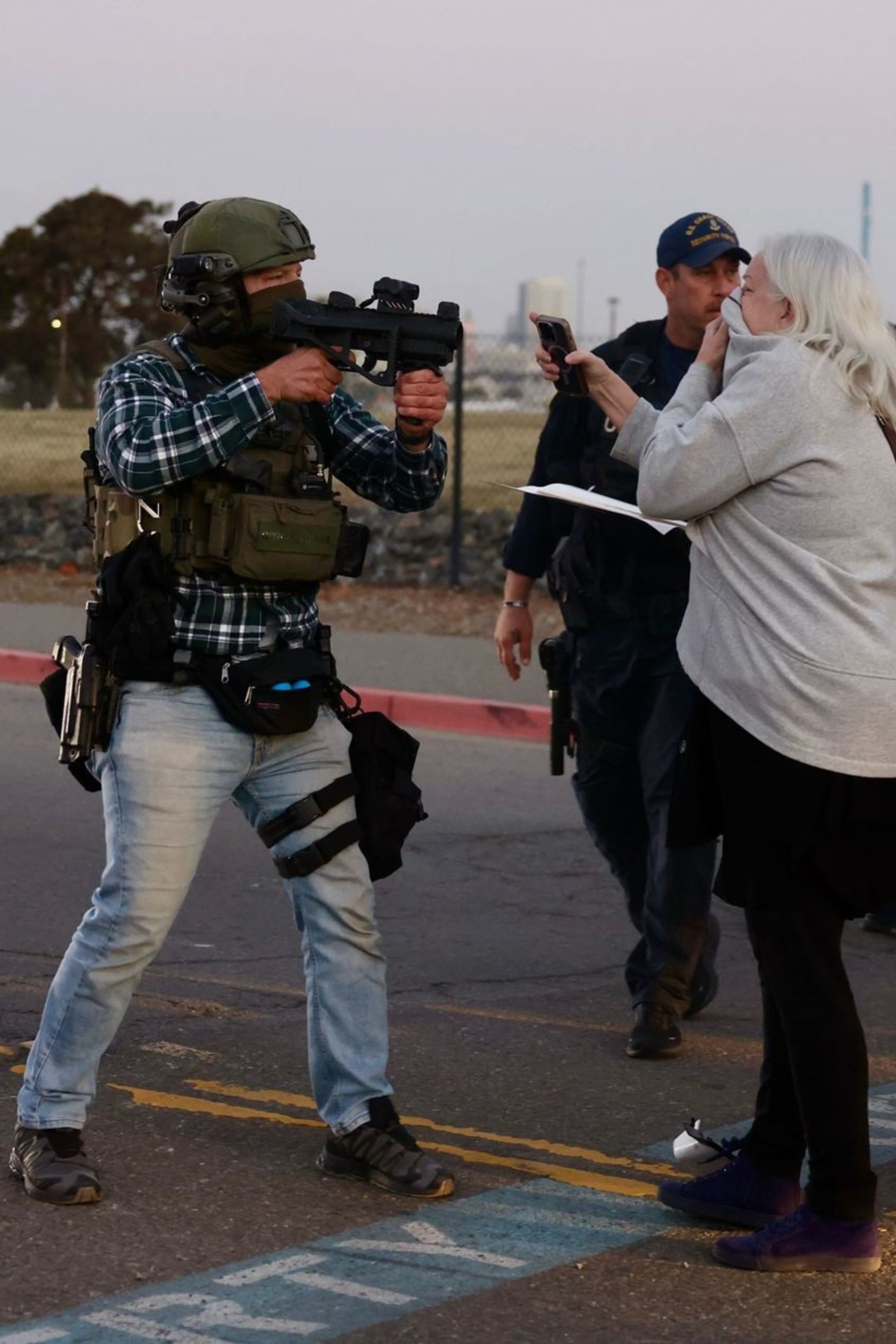 A man in tactical gear aims a gun at two women, one recording on her phone, while police officers approach in the background on a street.