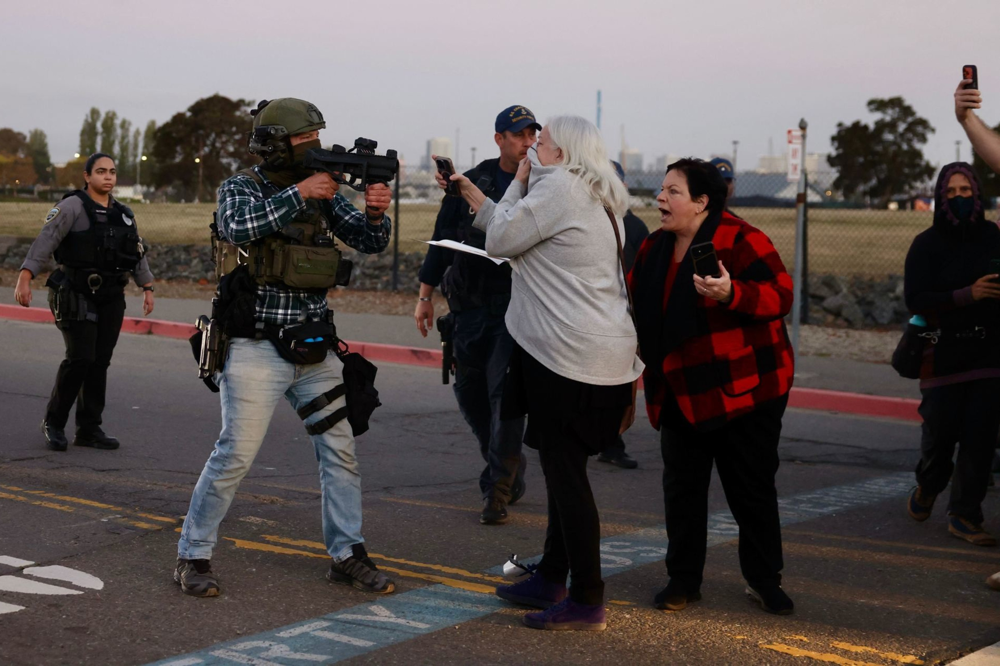 A man in tactical gear aims a gun at two women, one recording on her phone, while police officers approach in the background on a street.