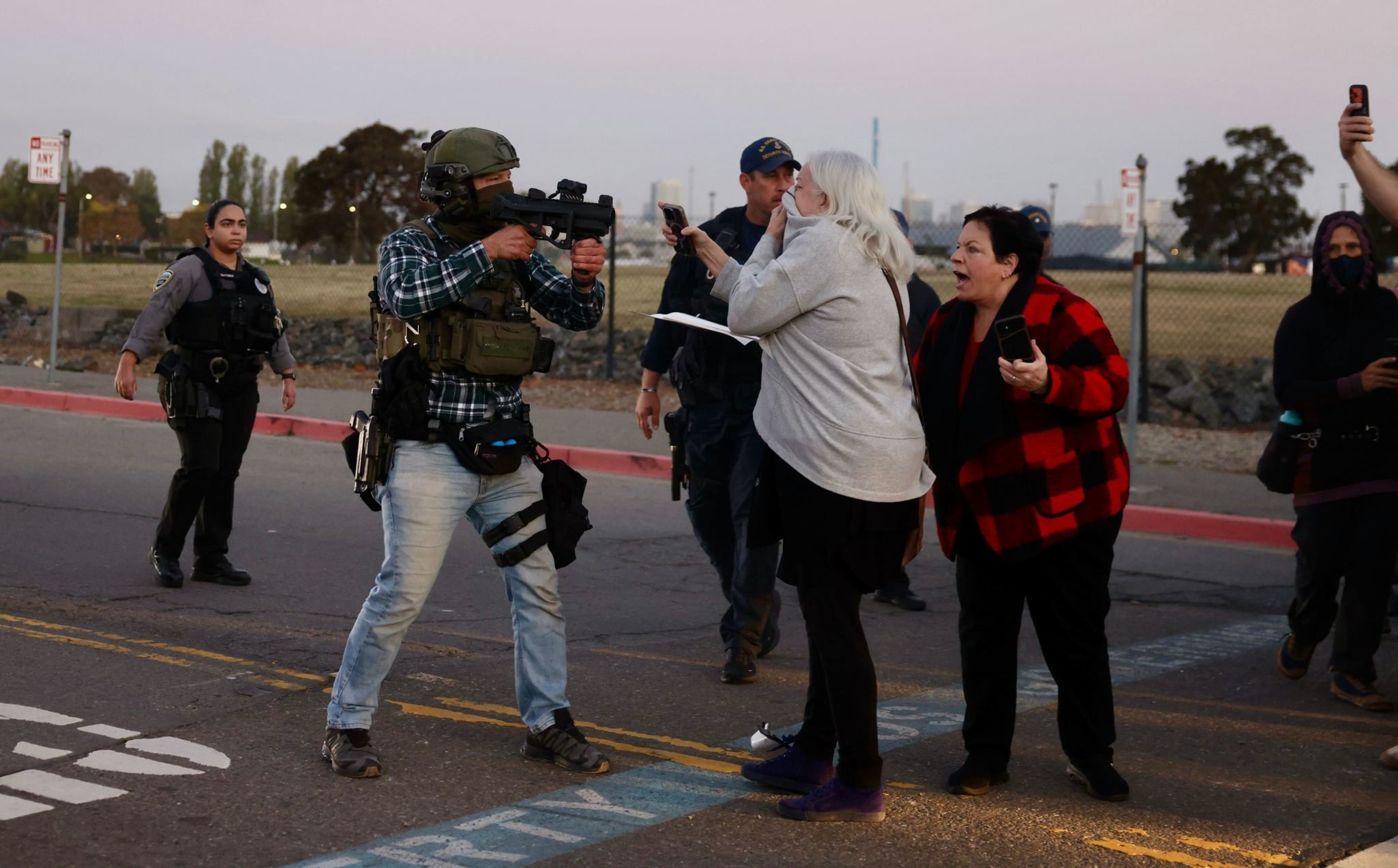 A man in tactical gear aims a gun at two women, one recording on her phone, while police officers approach in the background on a street.
