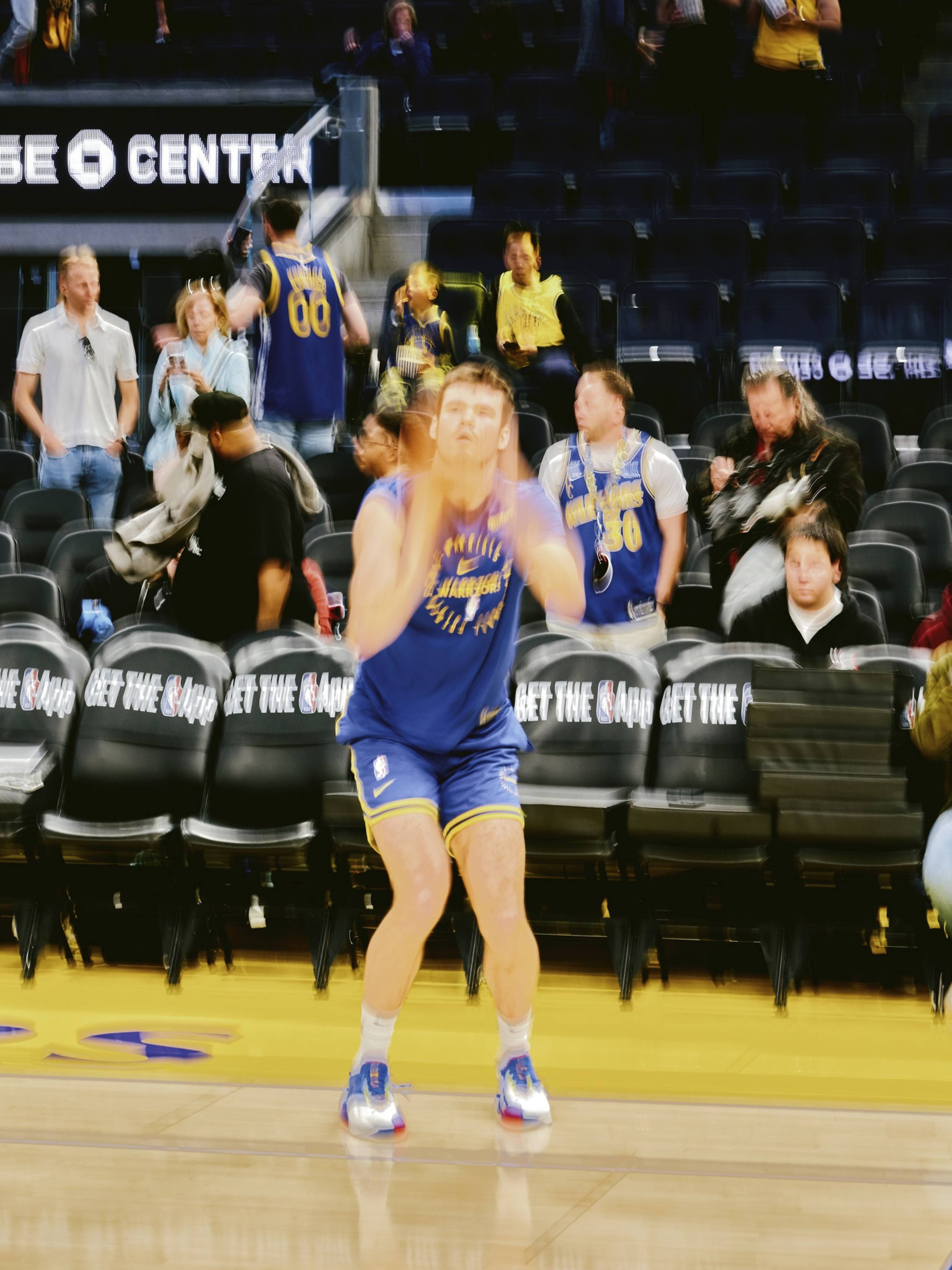 A basketball player in a blue and yellow uniform is preparing to shoot on the court, with blurred spectators and empty seats in the background.