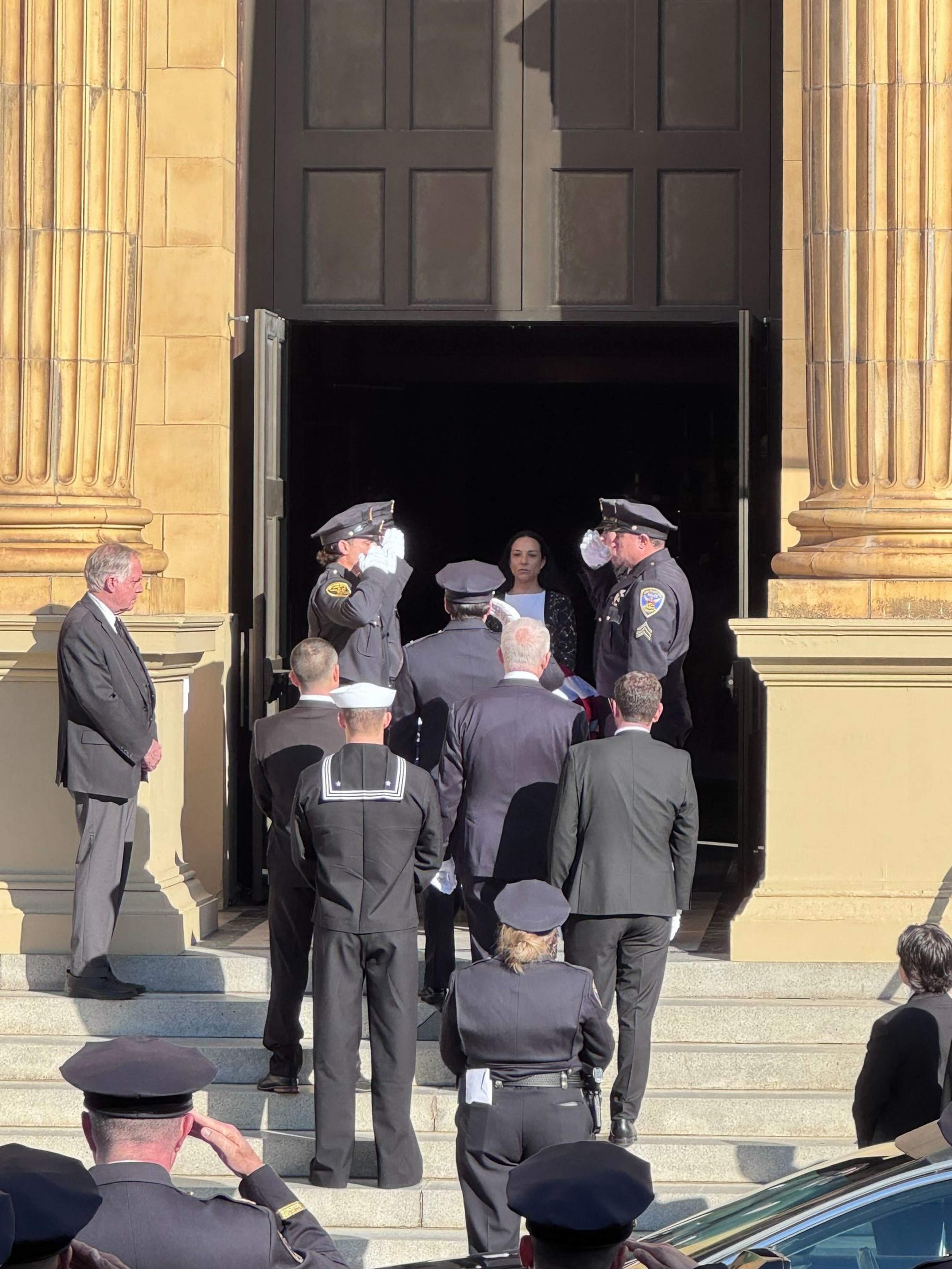 Uniformed officers and formally dressed people stand on steps outside a large building with columns, some saluting near an open dark doorway.