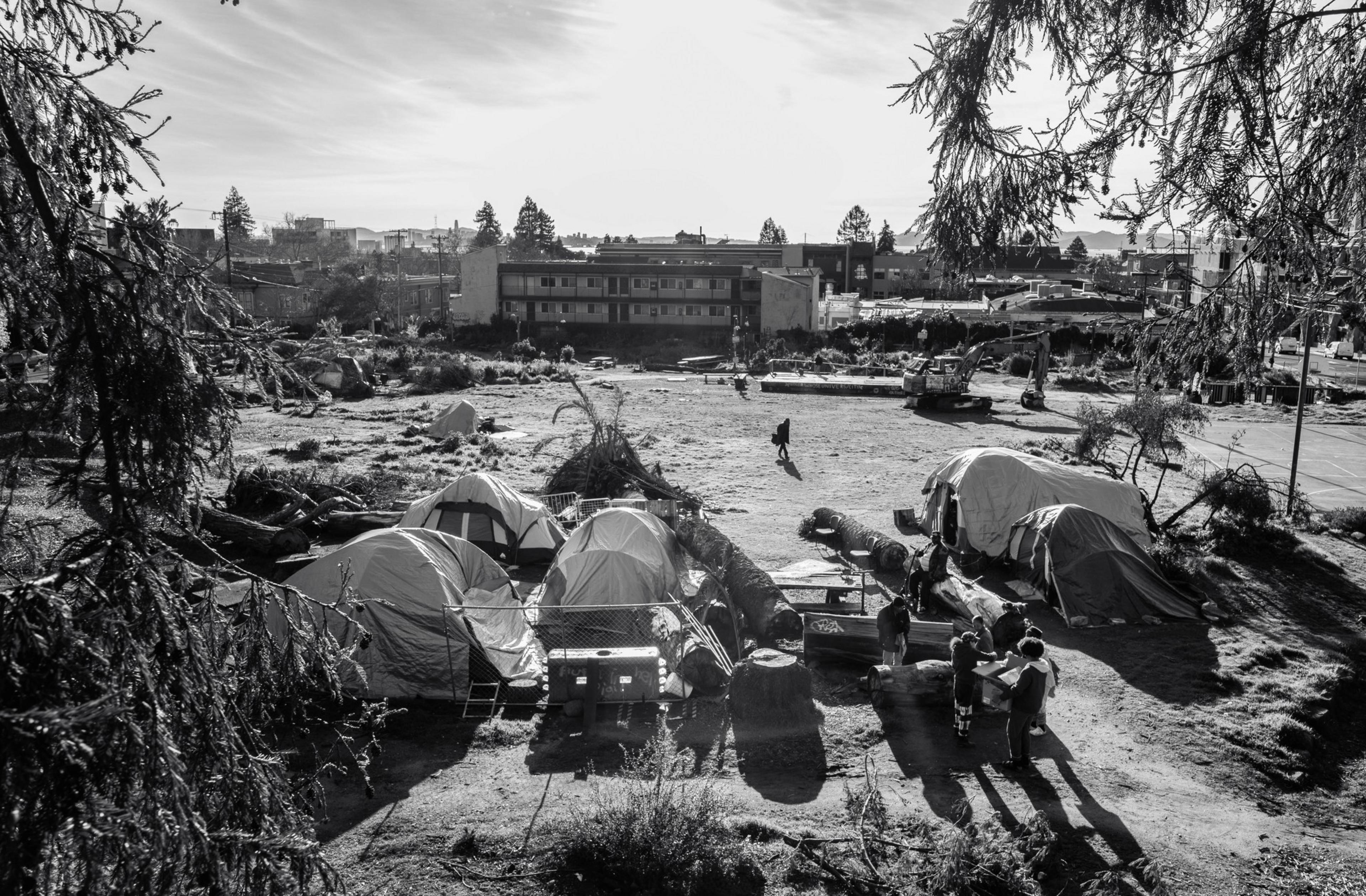 Several tents and makeshift shelters are set up in an open area surrounded by trees and buildings. People are gathered and talking near the tents.