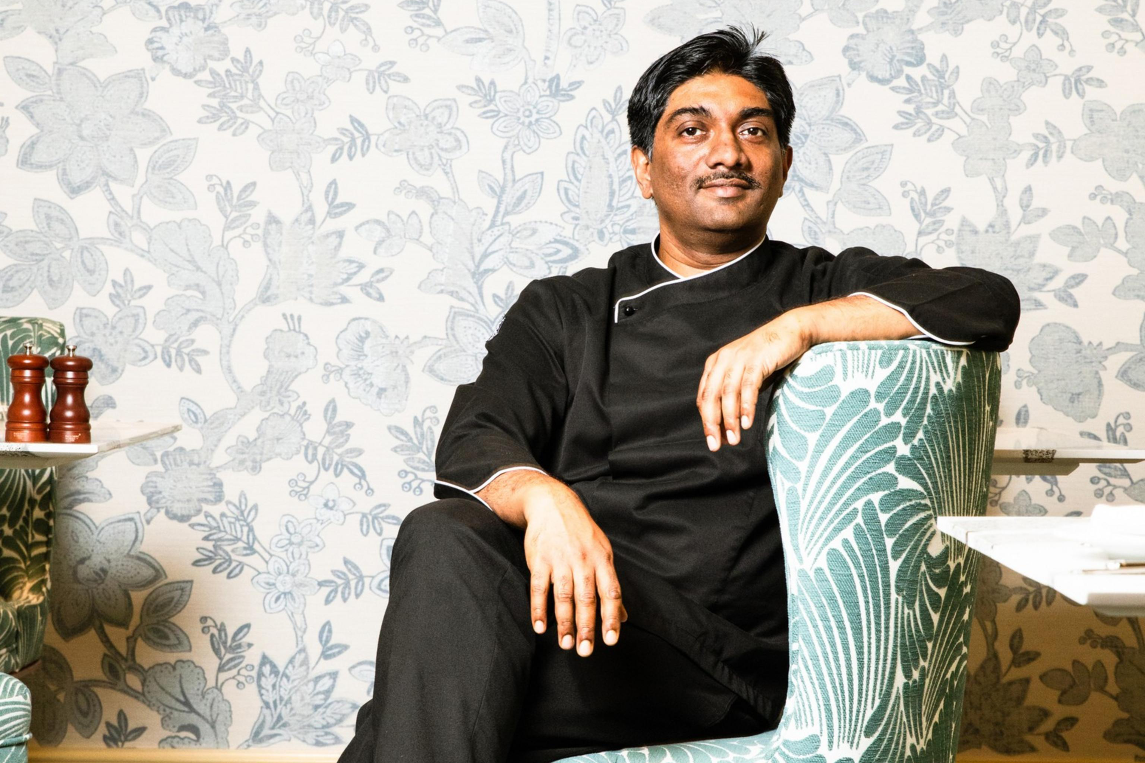 A man in black attire sits relaxed in a green-patterned armchair against a floral wallpaper backdrop, with his legs crossed and a confident expression.