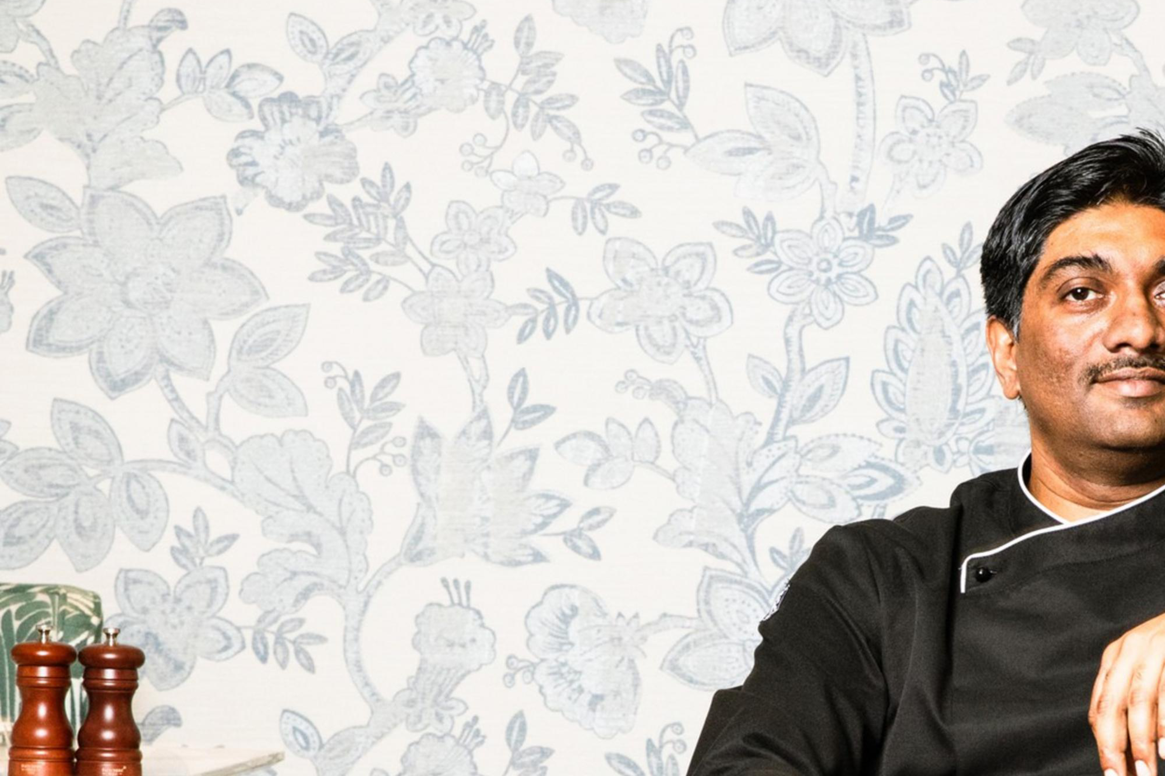 A man in black attire sits relaxed in a green-patterned armchair against a floral wallpaper backdrop, with his legs crossed and a confident expression.