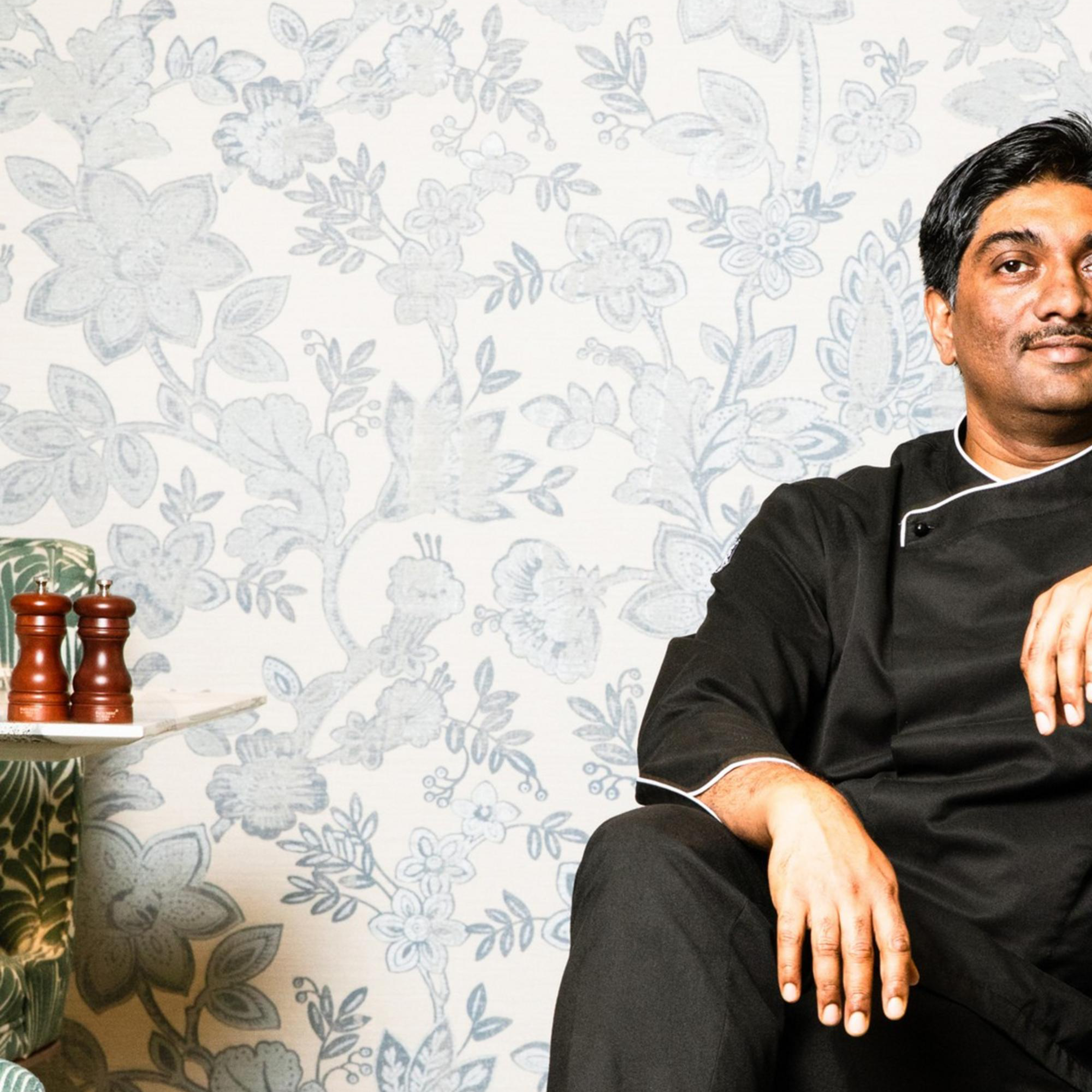 A man in black attire sits relaxed in a green-patterned armchair against a floral wallpaper backdrop, with his legs crossed and a confident expression.