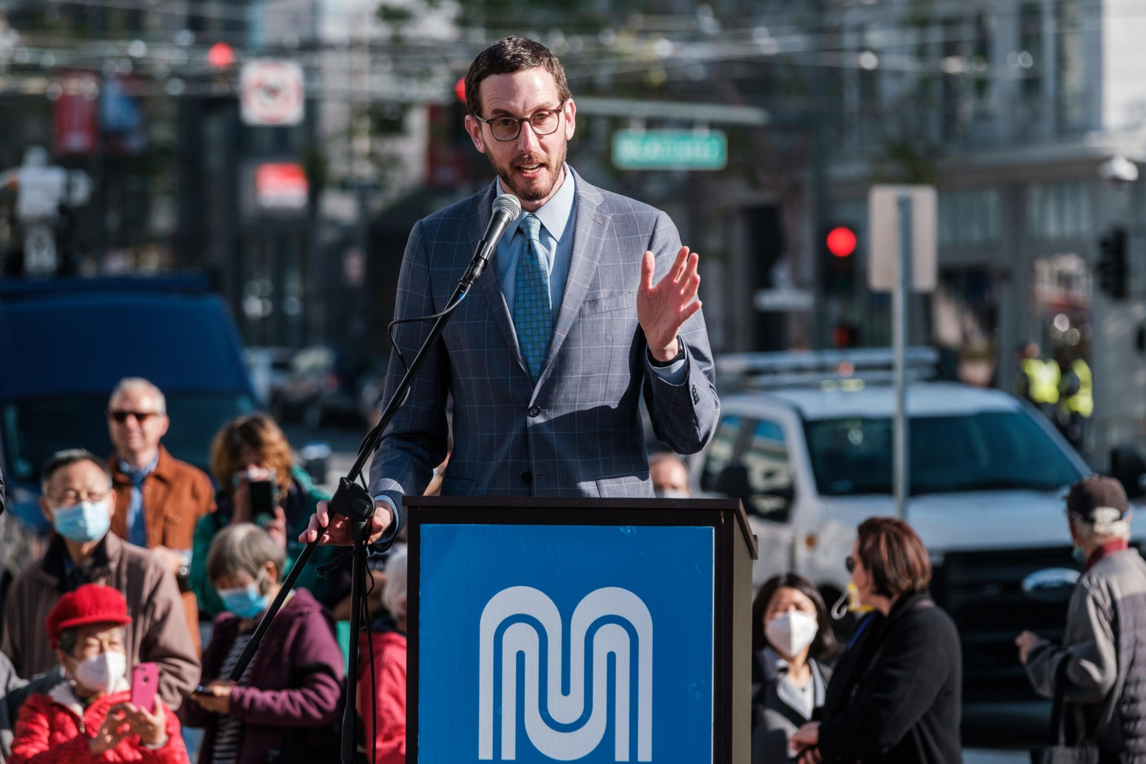 A man in a suit speaks at a podium with a blue emblem while people, some wearing masks, listen behind him on a city street.