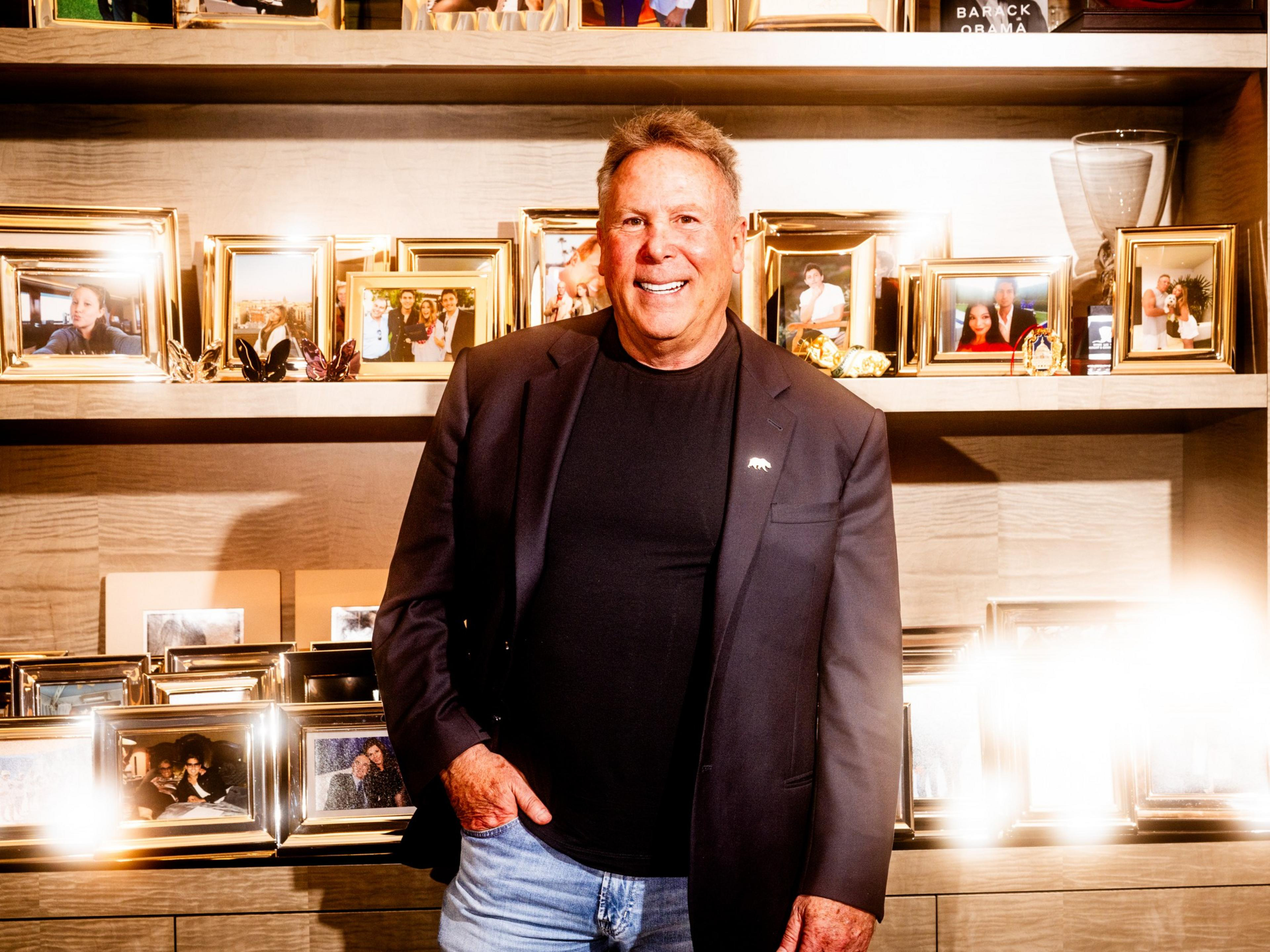 A man in a black blazer and jeans stands smiling in front of shelves filled with numerous framed family photos and decorative items.