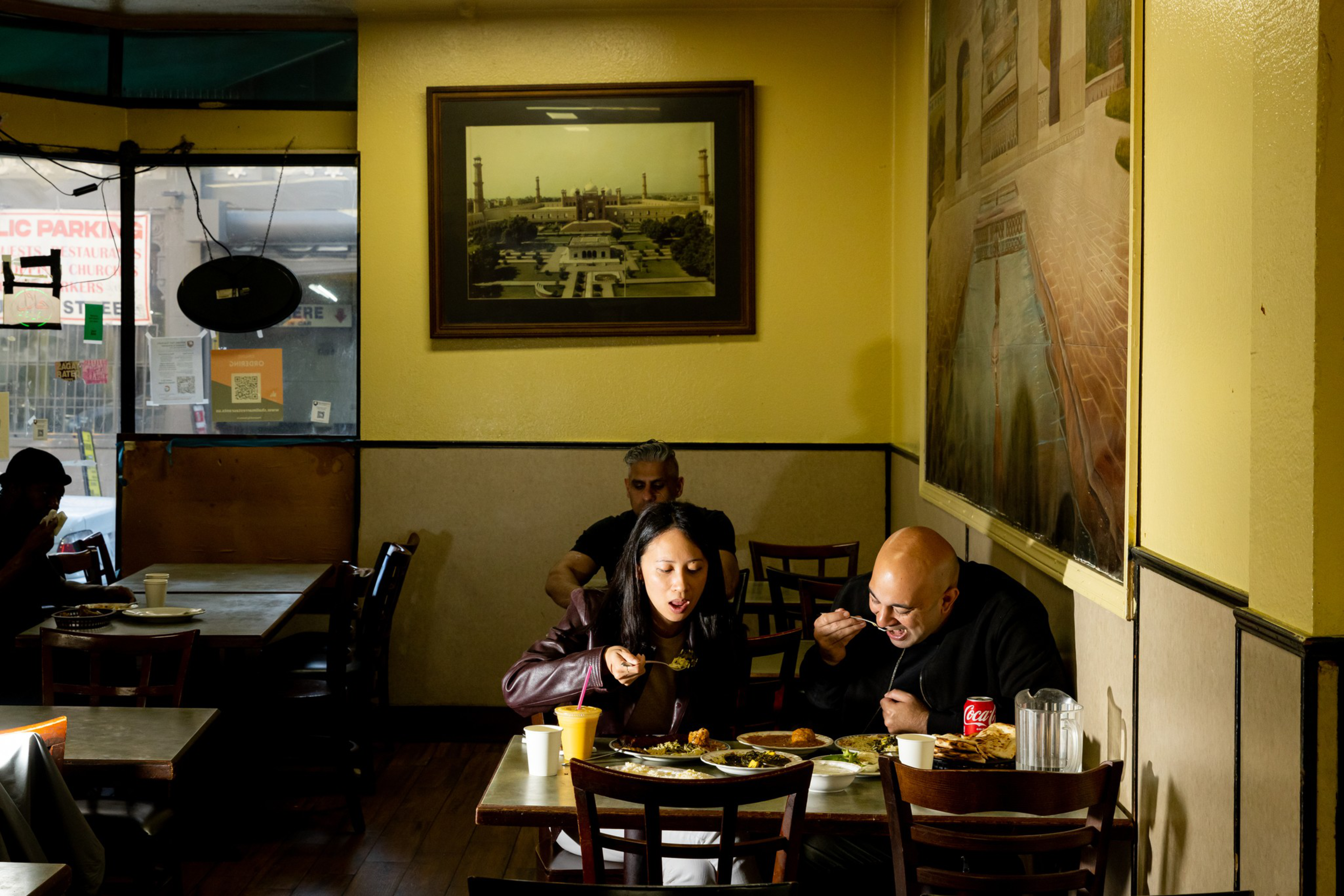 A woman and a bald man eat at a small restaurant table with plates of food, cups, and a Coca-Cola can, while a man sits alone in the background.