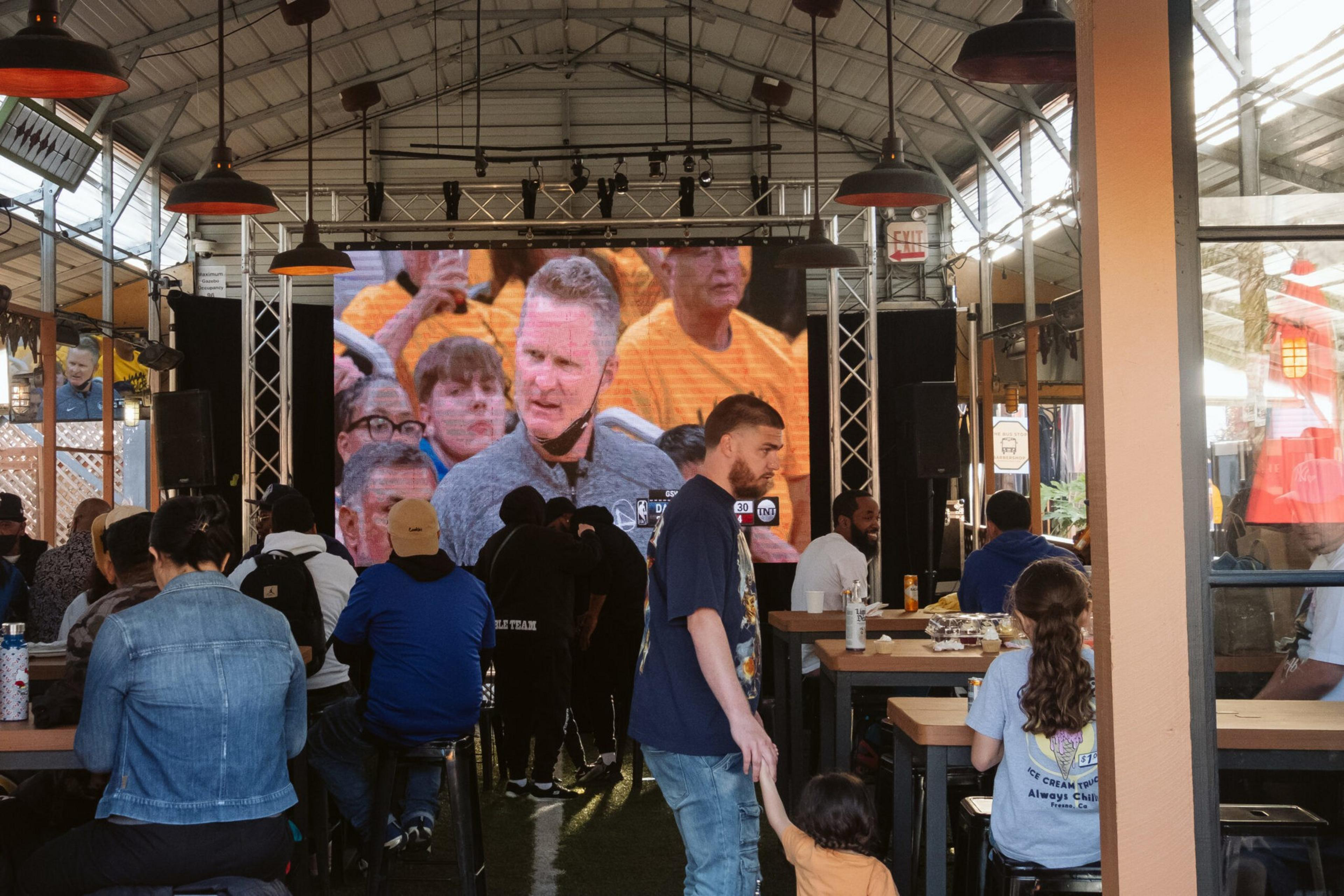 People sit and stand inside a covered outdoor space, watching a large screen showing a sports broadcast with a man speaking.