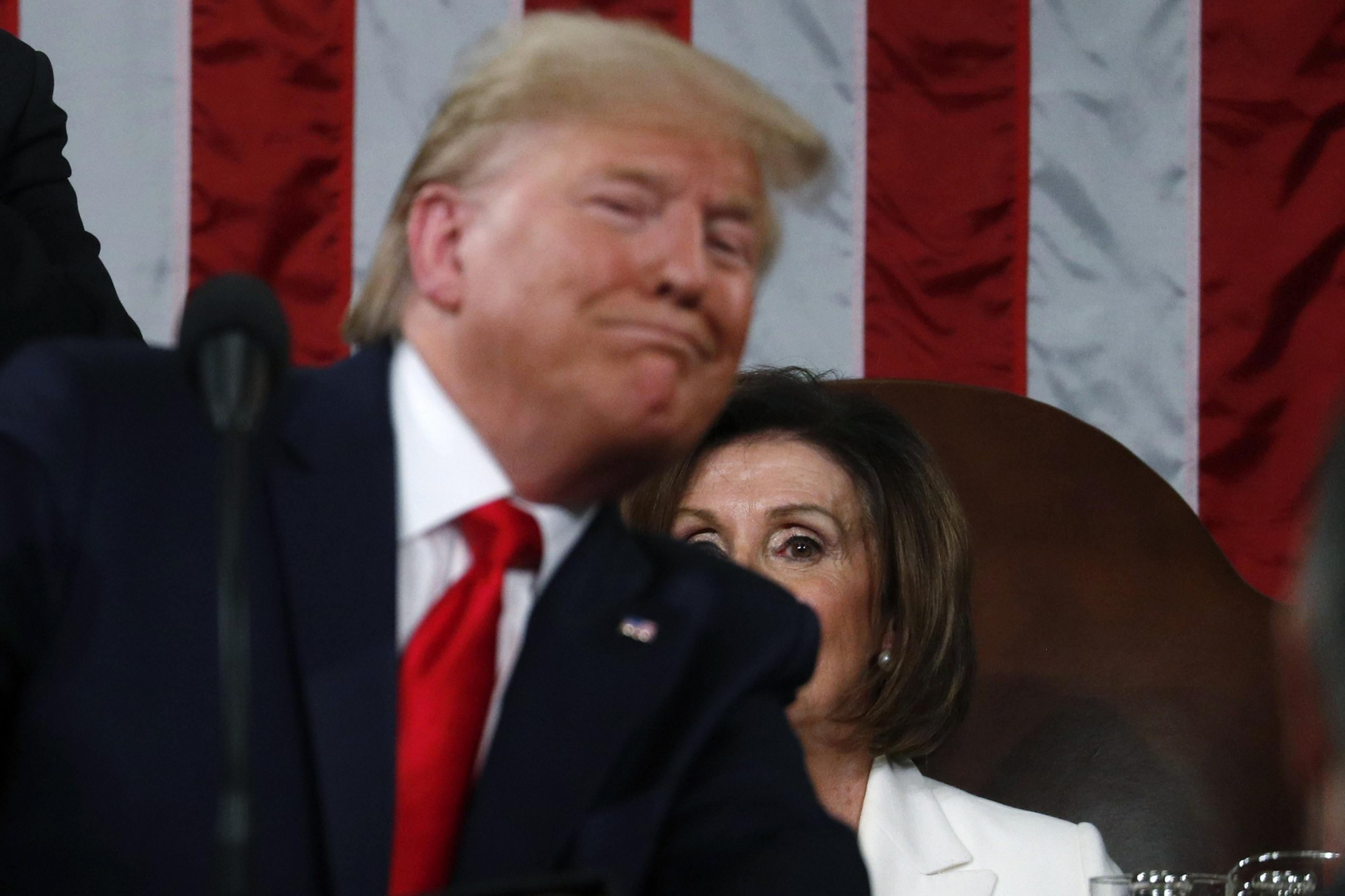 A person in a suit with a red tie is in the foreground, looking pleased. Behind them, another person in a white outfit is seated, with a serious expression. An American flag hangs in the background.