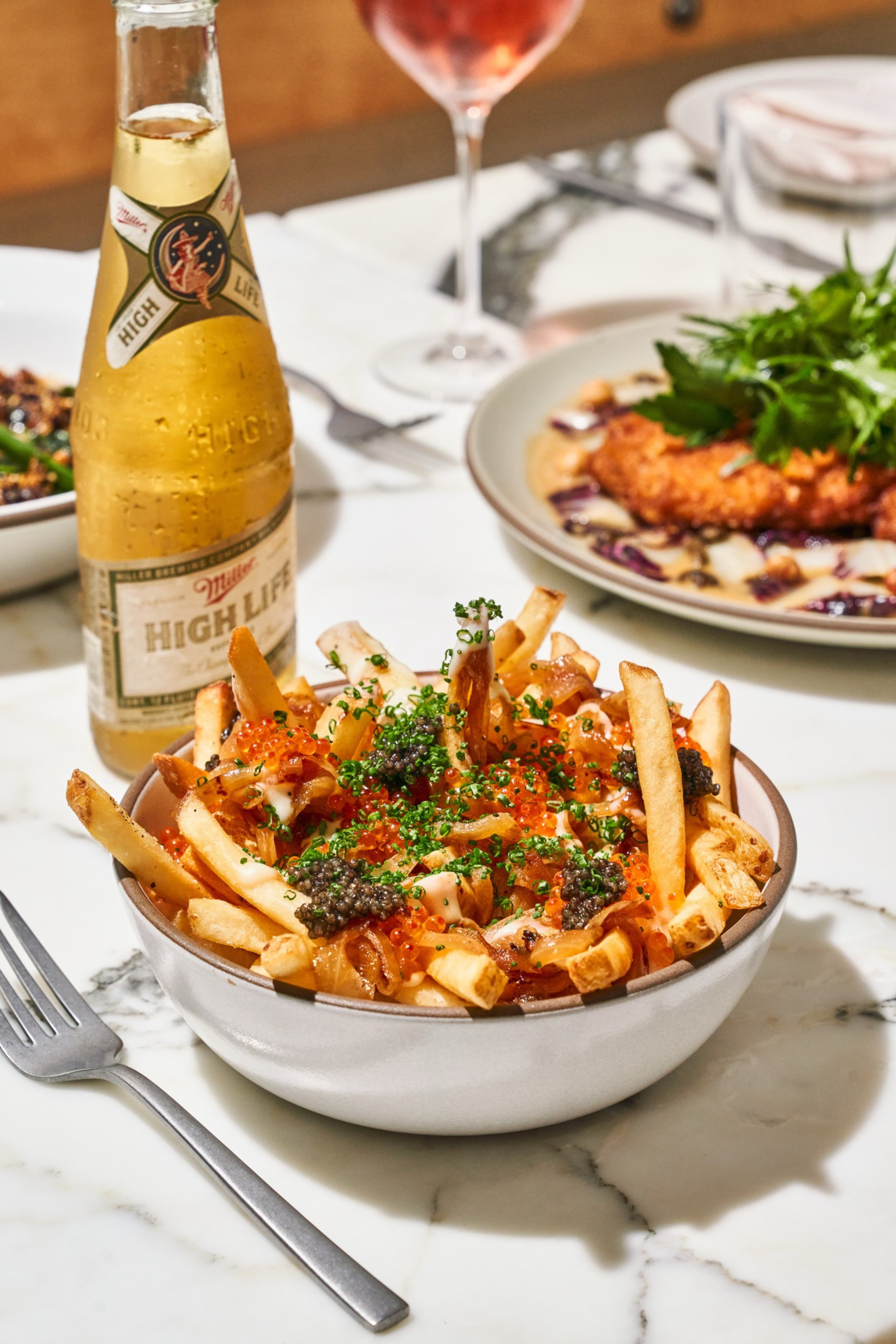 A bowl of fries topped with caviar, herbs, and sauce sits beside a bottle of beer on a marble table, with a plated dish and wine glass in the background.
