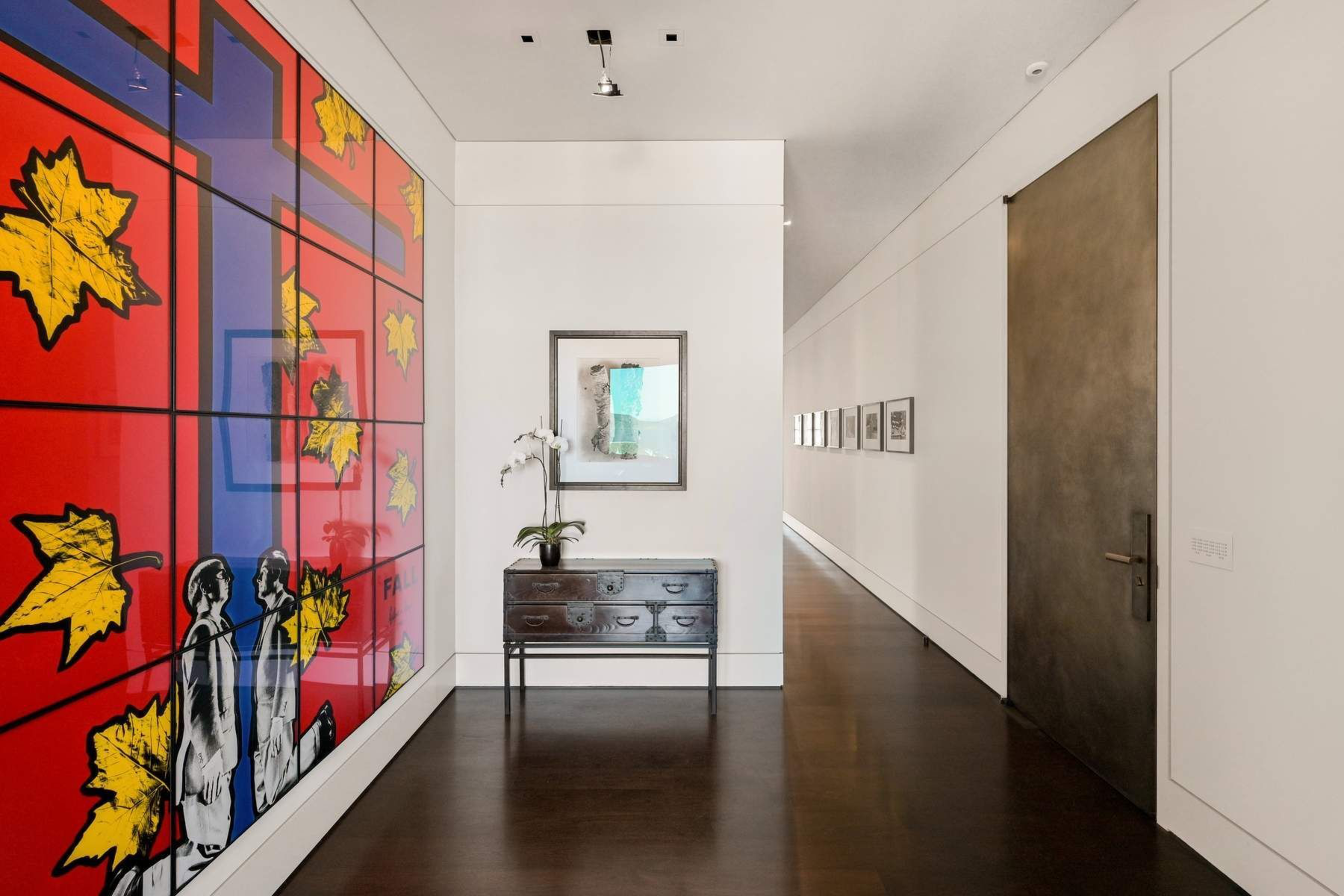 A modern hallway with dark wood floors features a large art piece with yellow leaves on red and blue, a small chest with an orchid, and framed pictures on white walls.
