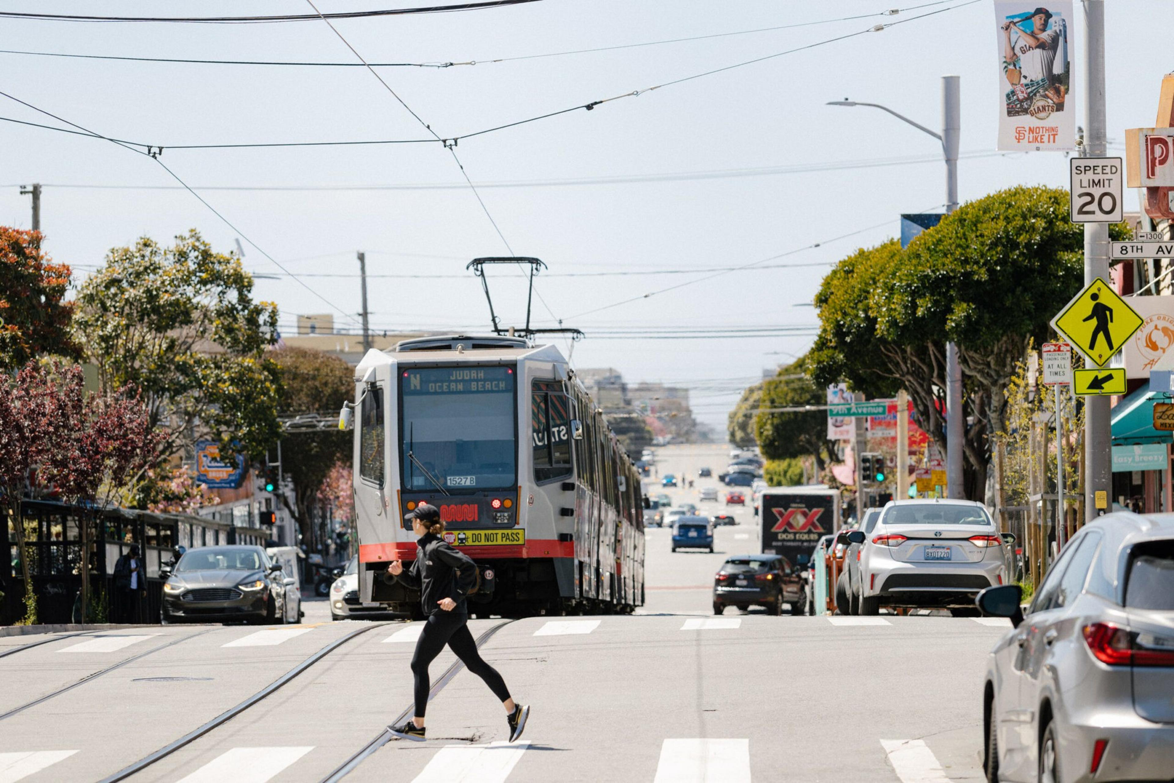 A person in black athletic wear crosses a street at a crosswalk while a city light rail train approaches from behind, with cars and trees lining the street.