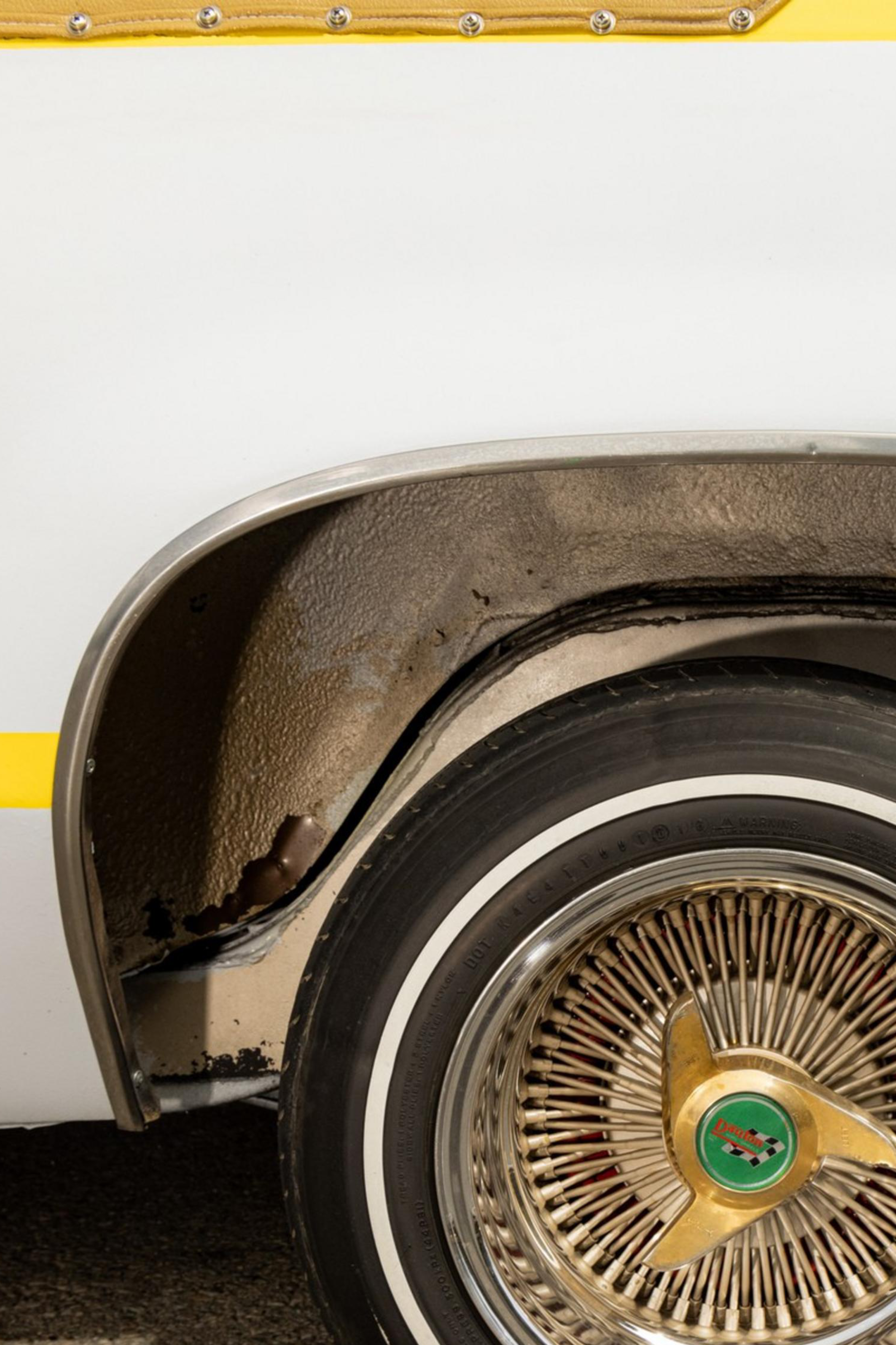 The image shows a close-up of a vintage car's rear wheel with a gold wire-spoke rim, whitewall tire, and a white and yellow paint job. The car's wheel well appears slightly rusted.