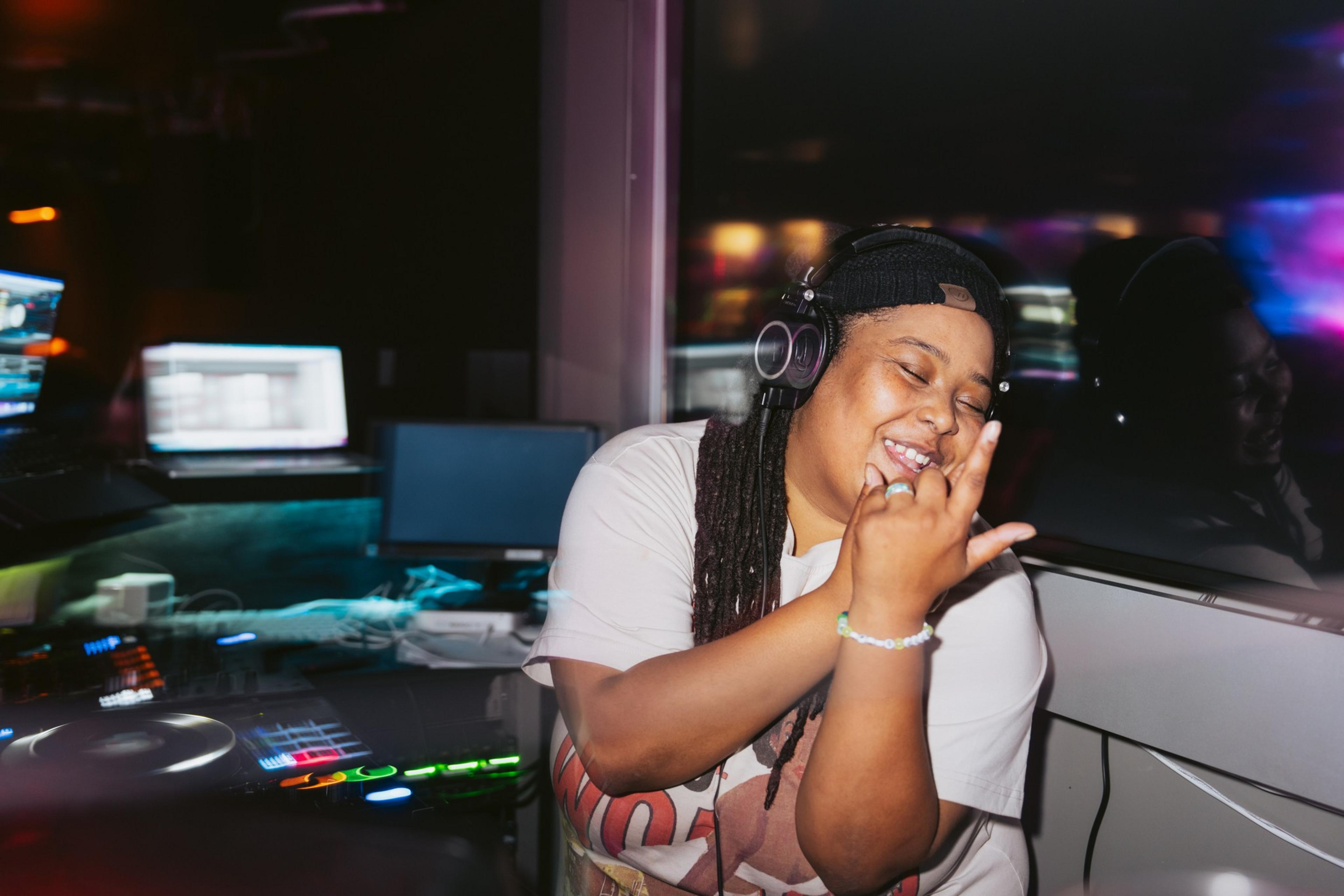 A smiling DJ, wearing headphones and a white shirt, is in a dimly lit room with computer screens and colorful lights around the DJ setup.