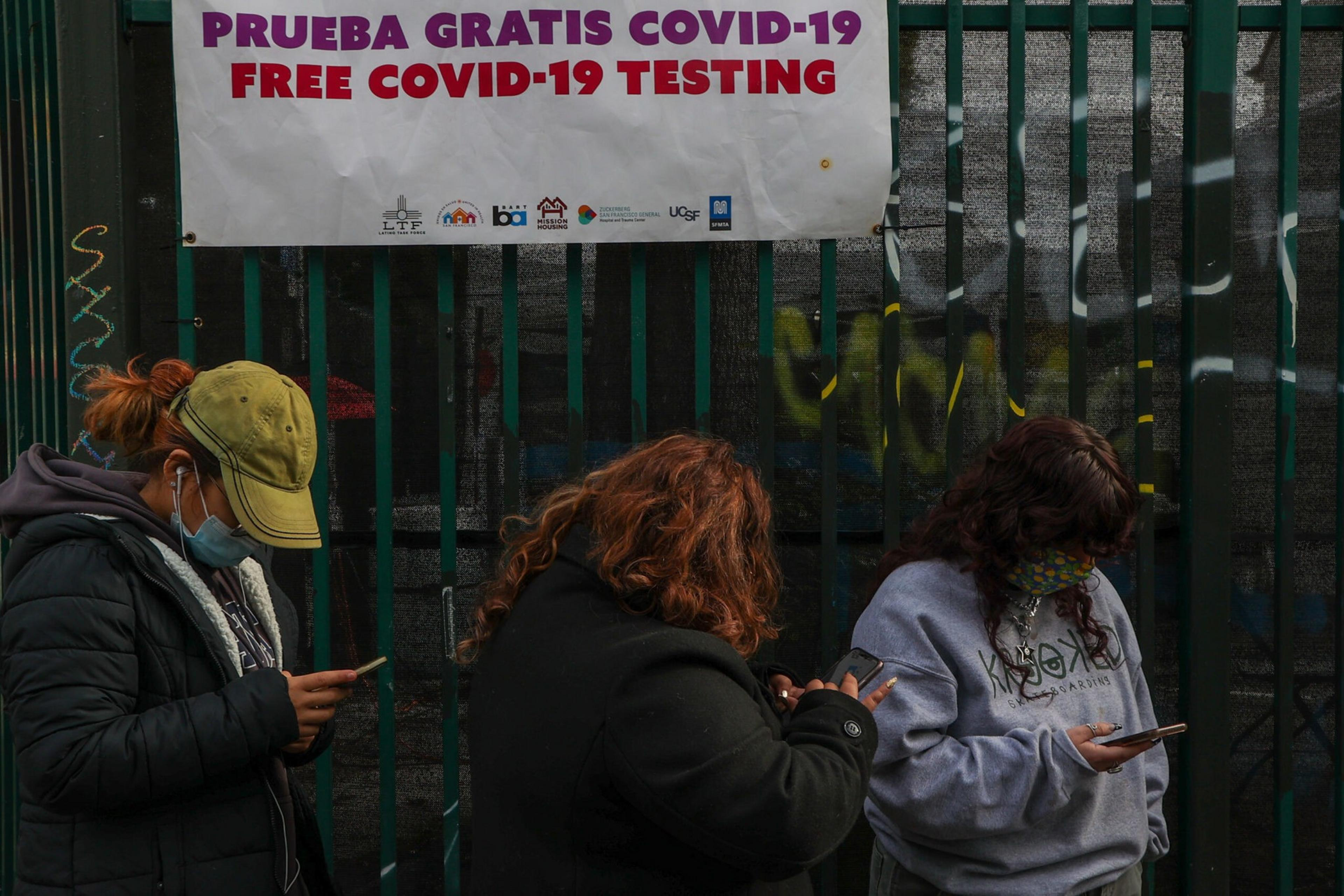 Three people wearing masks stand in a line looking at their phones under a sign offering free COVID-19 testing in Spanish and English.