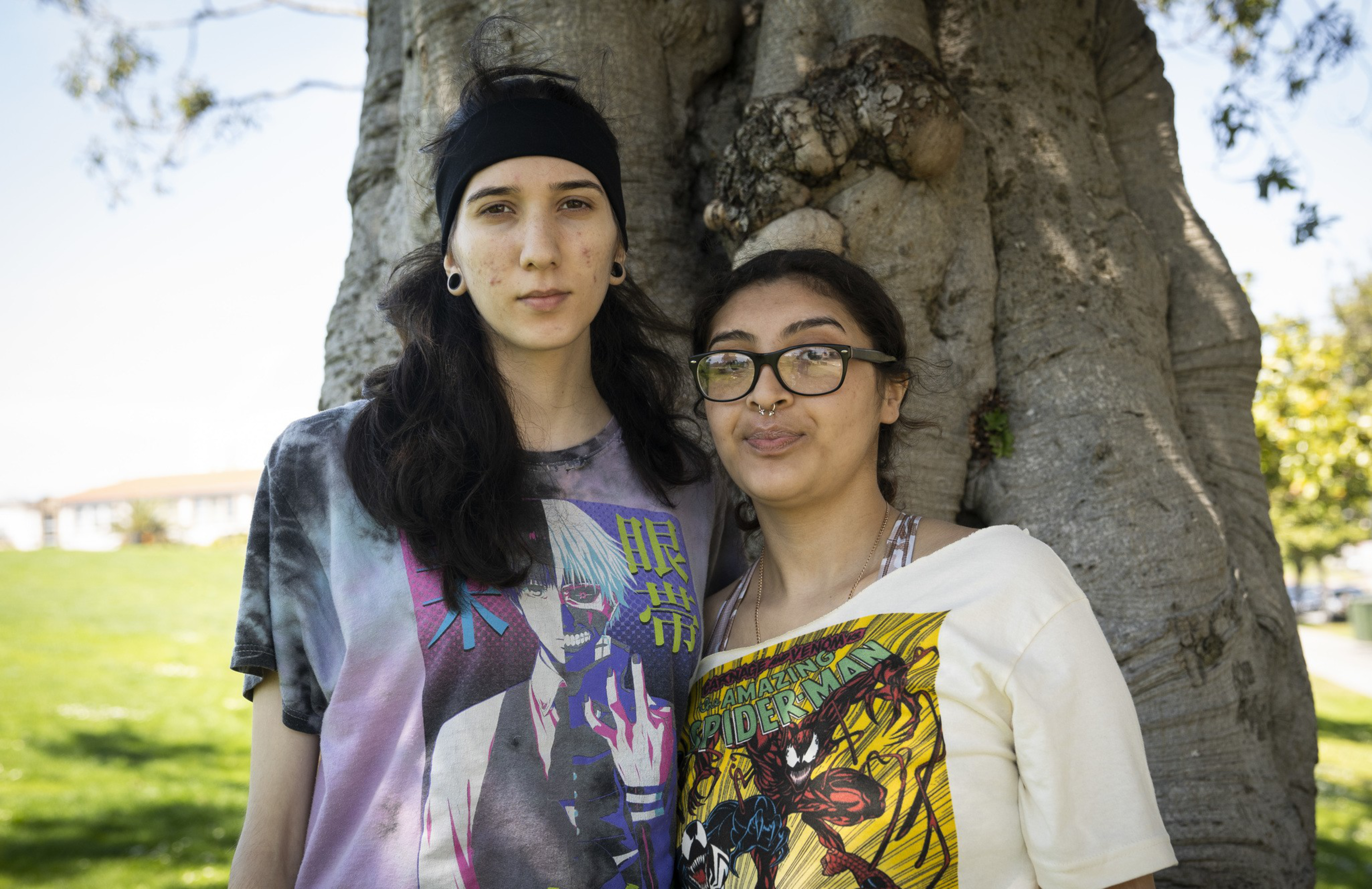 Two people stand in front of a large tree, wearing graphic t-shirts with anime and comic designs. They appear relaxed, with greenery in the background.
