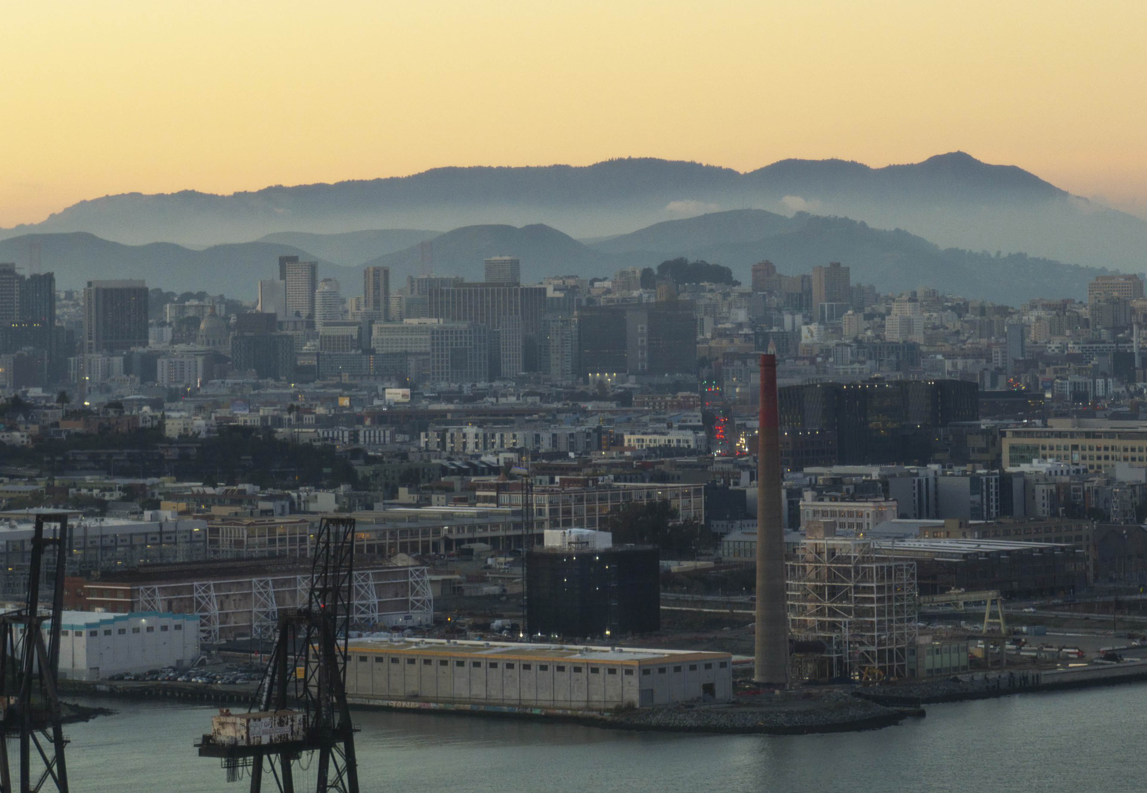 A city skyline at sunset with industrial buildings and cranes in the foreground, water along the bottom, and misty mountains in the background.