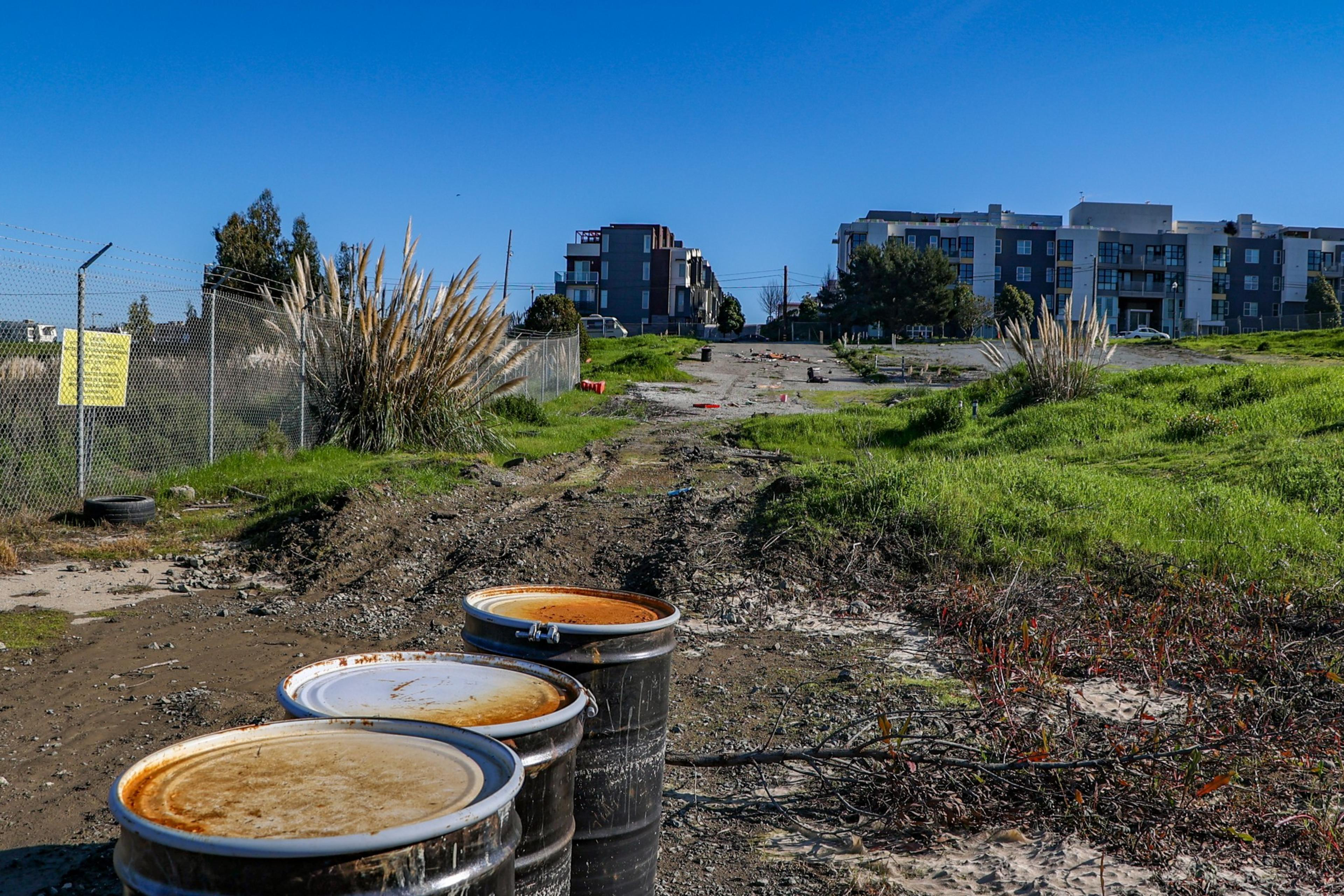 Three rusty barrels sit on a muddy path with scattered vegetation, a wire fence on the left, and distant modern buildings under a clear blue sky.