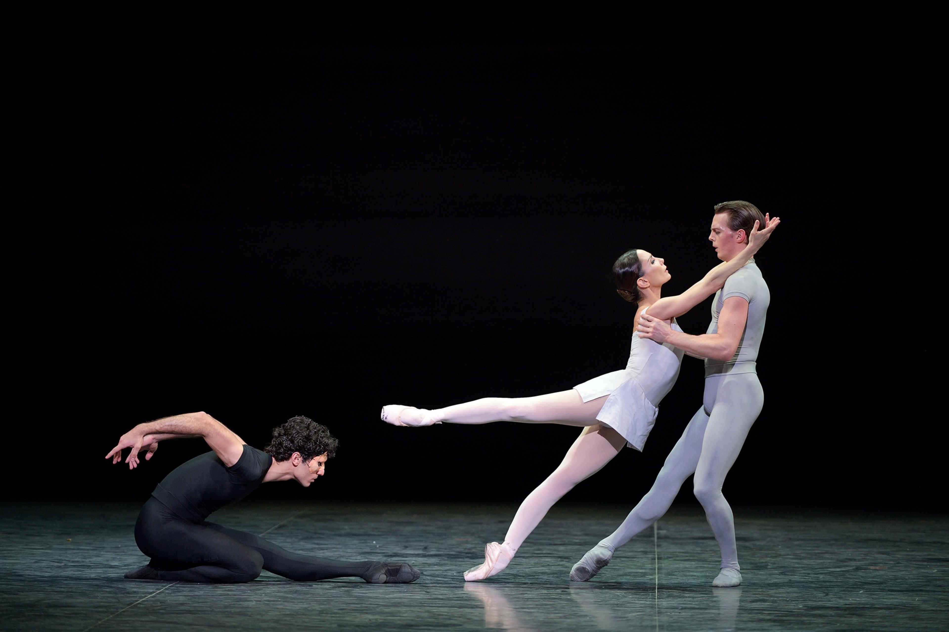 A male ballet dancer in black stretches low on the floor as a female dancer in white is lifted by a male dancer in gray against a dark background.