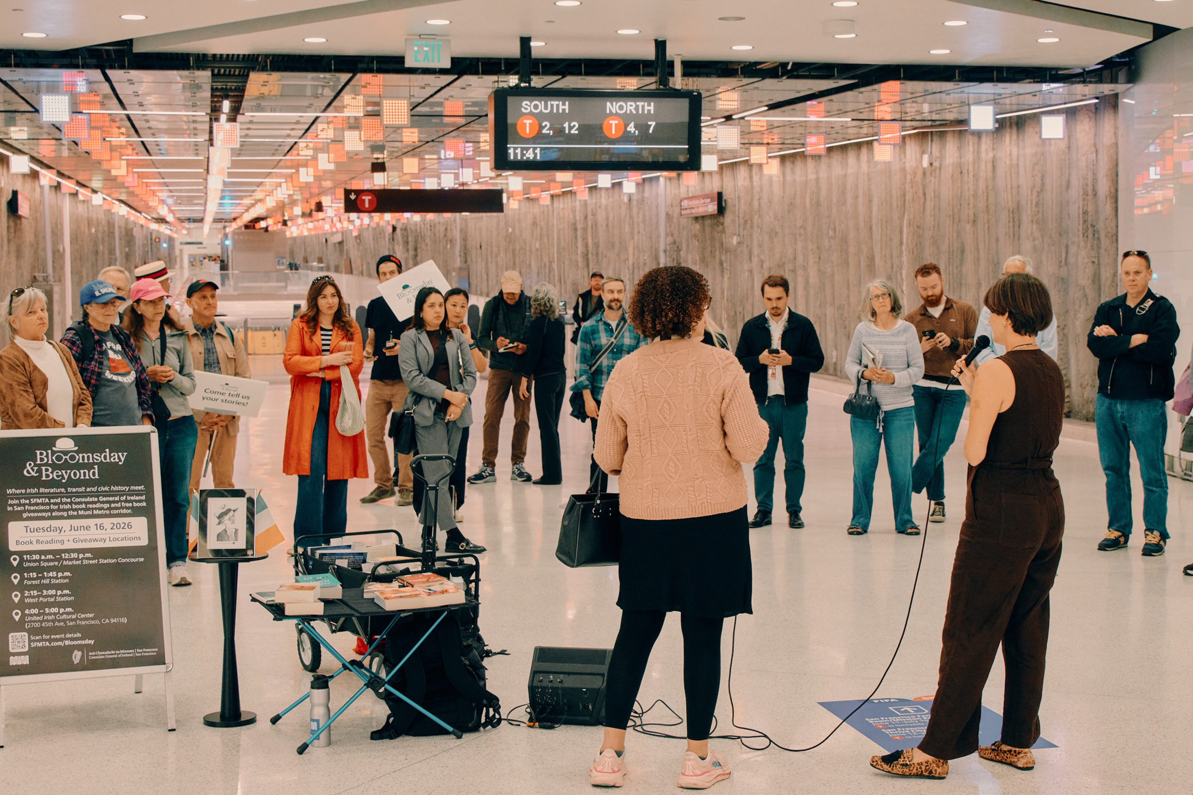 A group of people listens to two speakers in a subway station, with posters and equipment set up nearby. A digital sign shows train times.