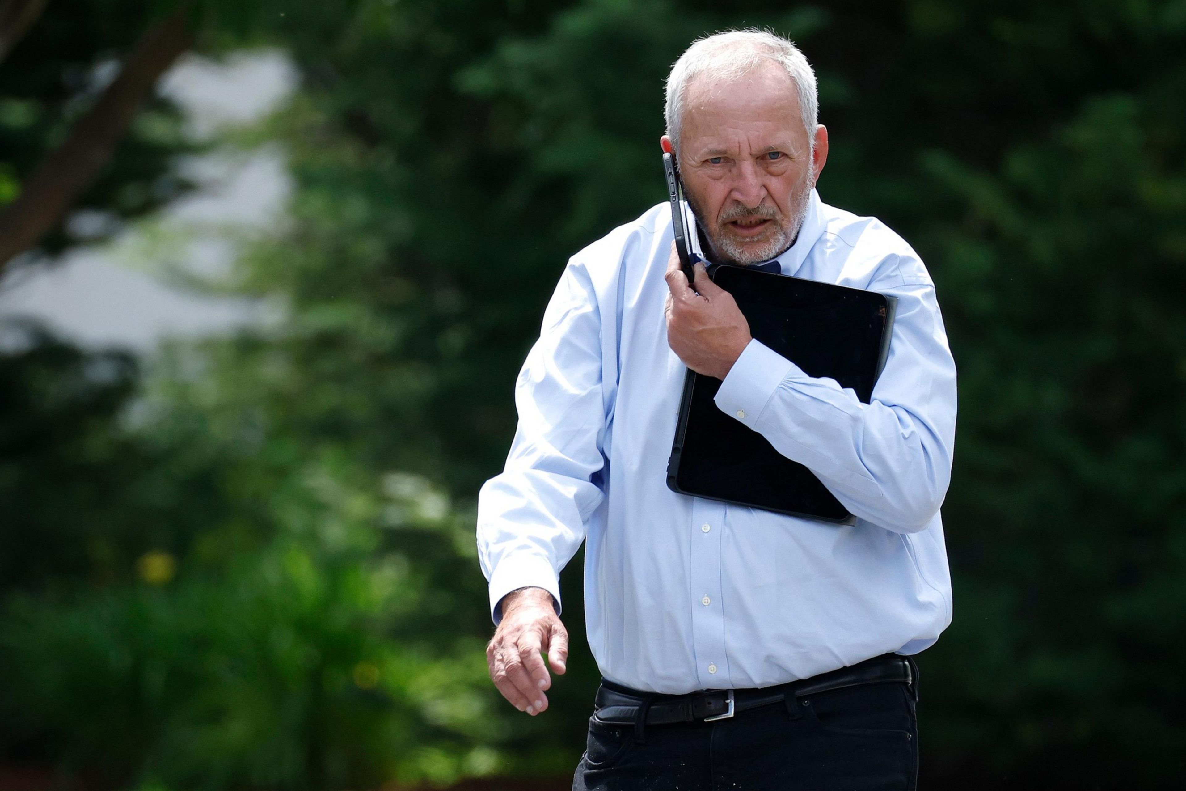 An older man with white hair and a beard walks outdoors, holding a laptop against his chest and a cell phone to his ear, wearing a light blue shirt and black pants.