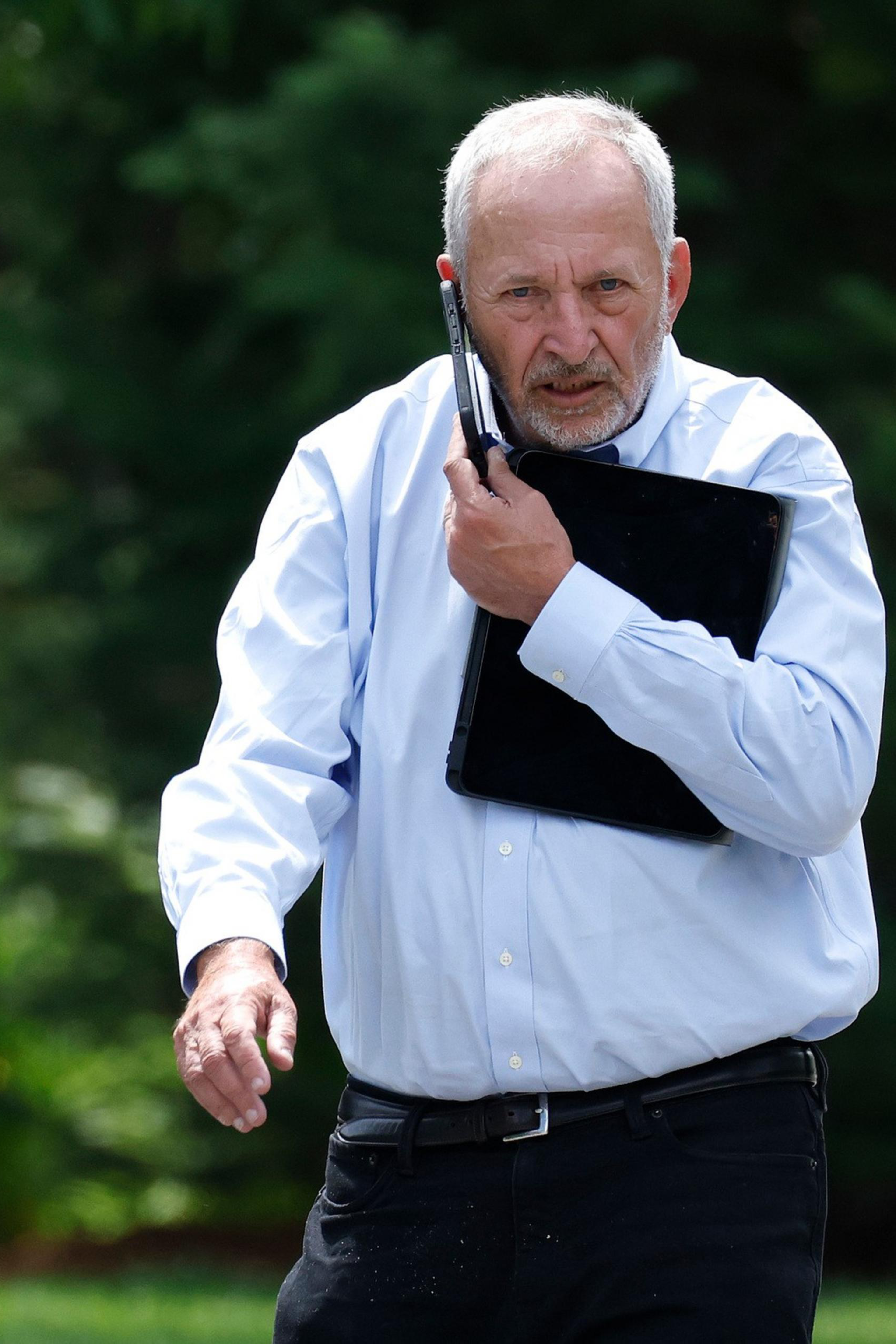 An older man with white hair and a beard walks outdoors, holding a laptop against his chest and a cell phone to his ear, wearing a light blue shirt and black pants.