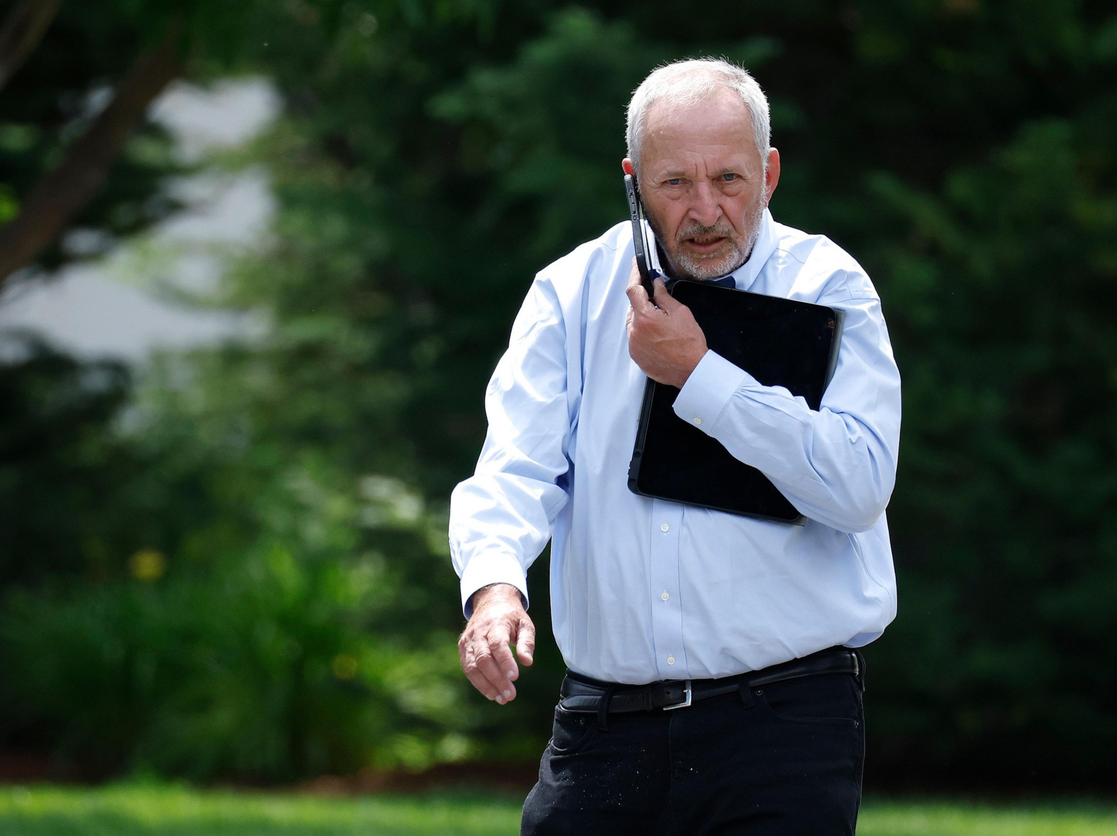 An older man with white hair and a beard walks outdoors, holding a laptop against his chest and a cell phone to his ear, wearing a light blue shirt and black pants.