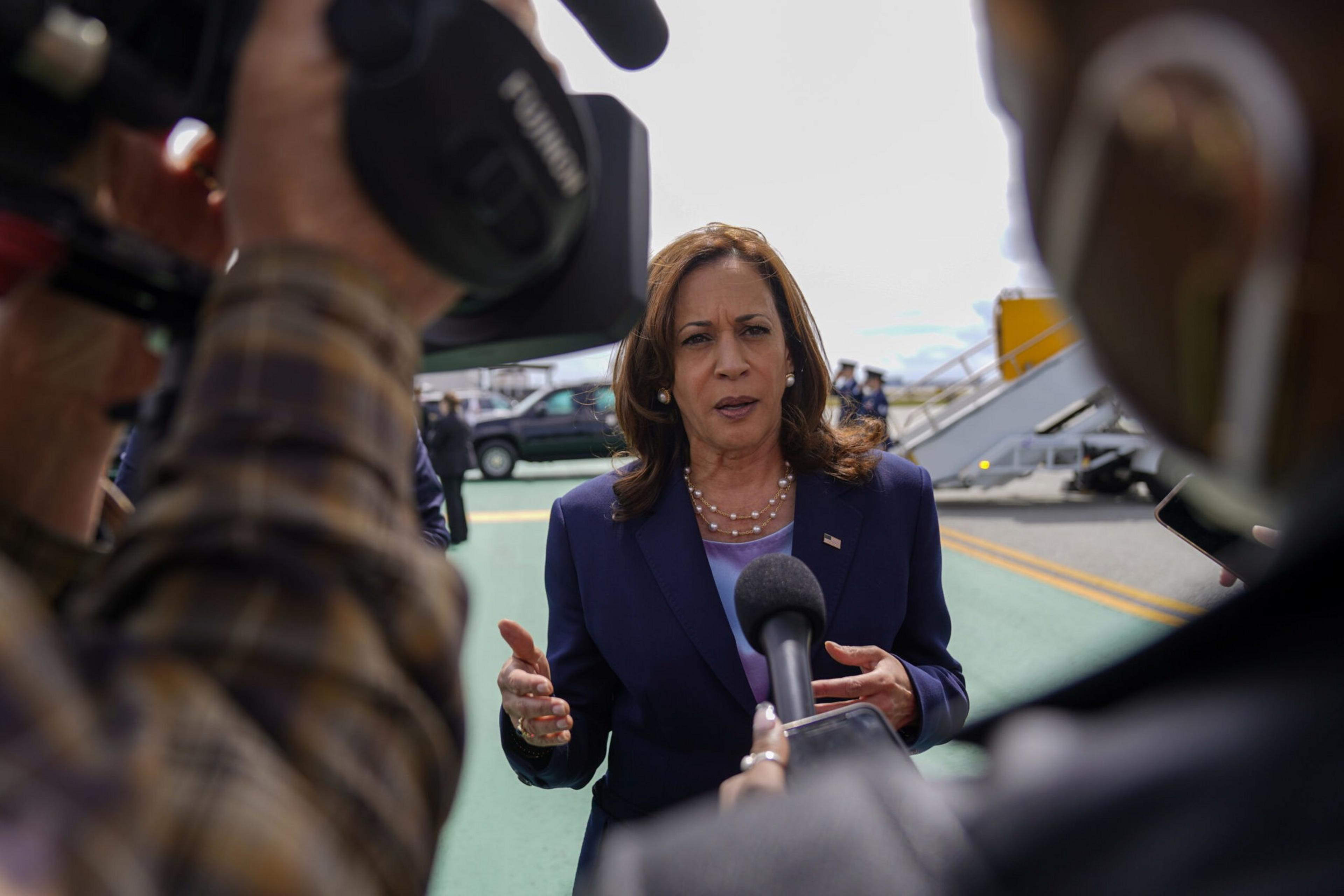A woman in a dark suit speaks to the press outdoors, flanked by microphones and cameras. In the background, a staircase leads to an elevated area.