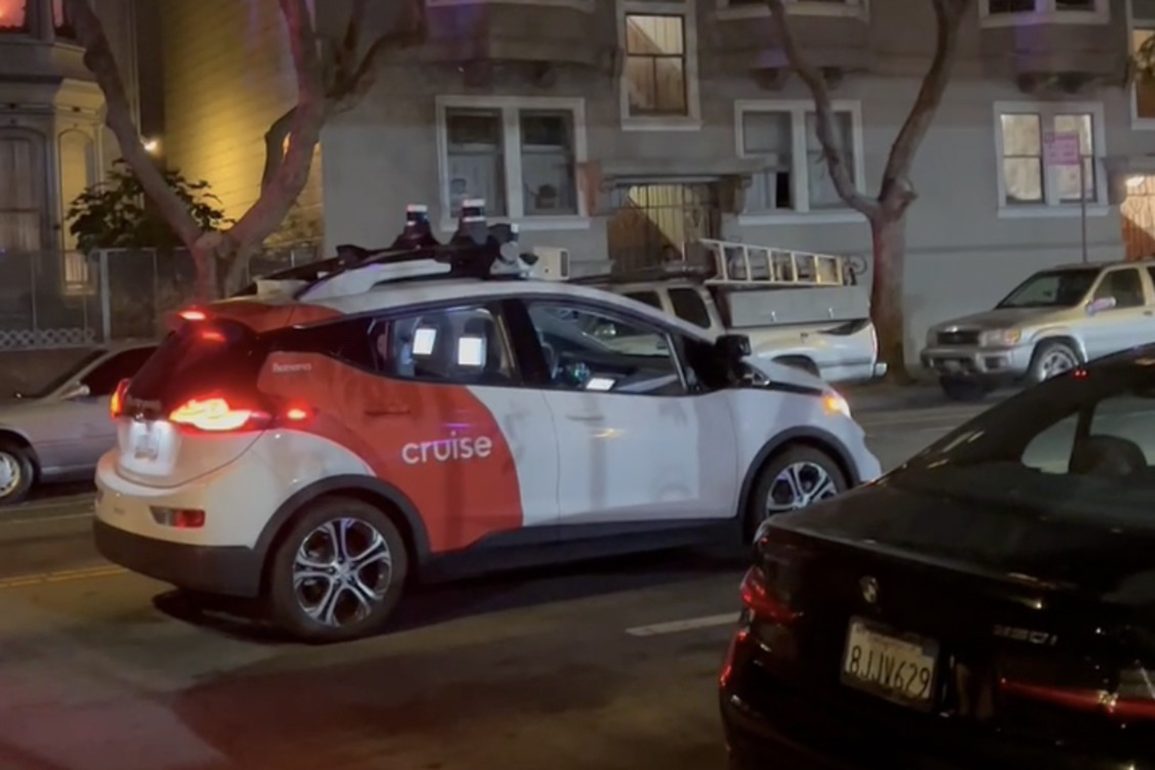 A white and orange autonomous Cruise vehicle is stopped on a city street at night with other parked cars and buildings visible.