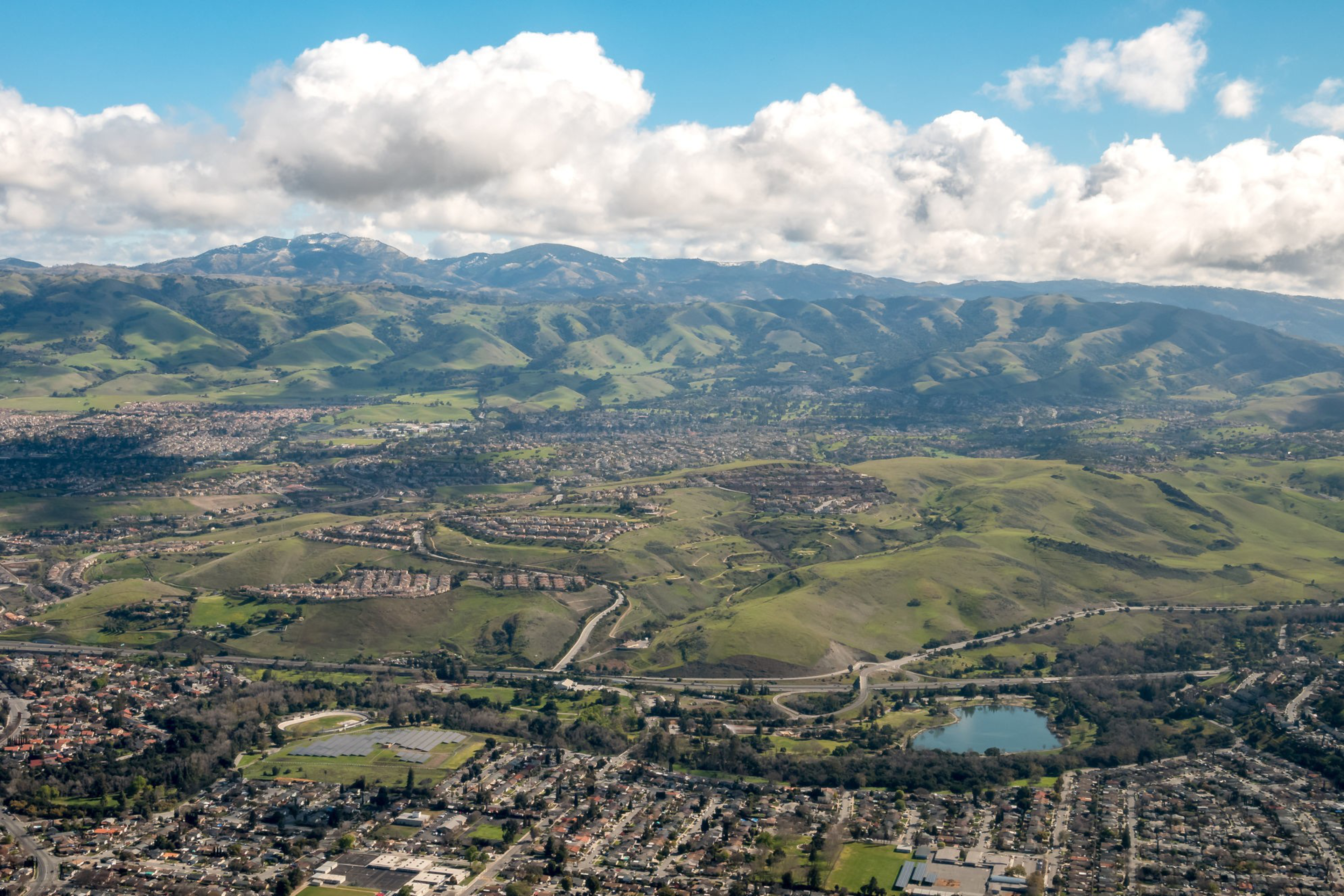 A sprawling urban area borders rolling green hills with a small lake, set against a backdrop of large, cloud-dotted mountains under a blue sky.