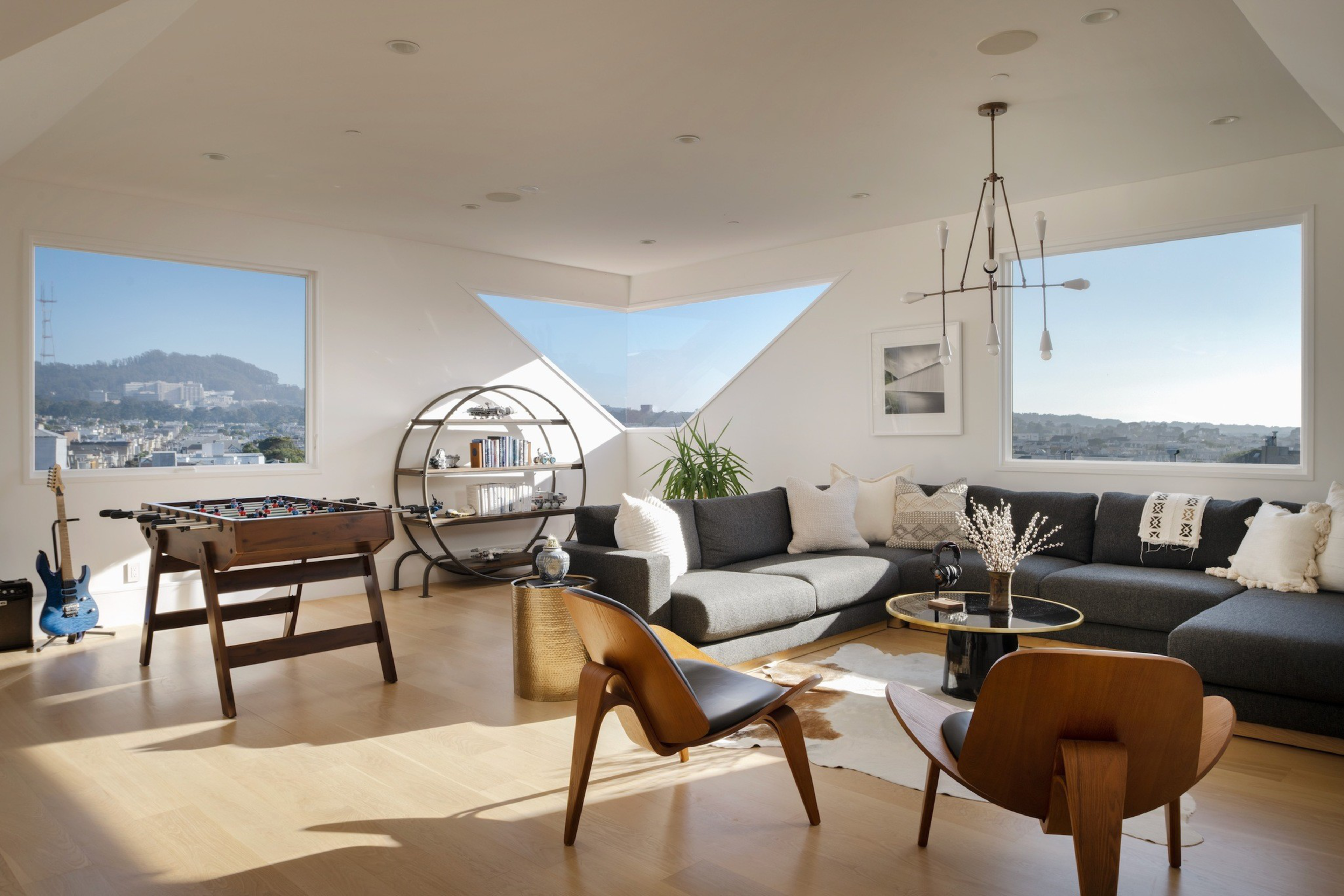 A bright living room with large windows, a gray sectional sofa, two wooden chairs, a foosball table, a round shelf, a blue guitar, and a modern chandelier.