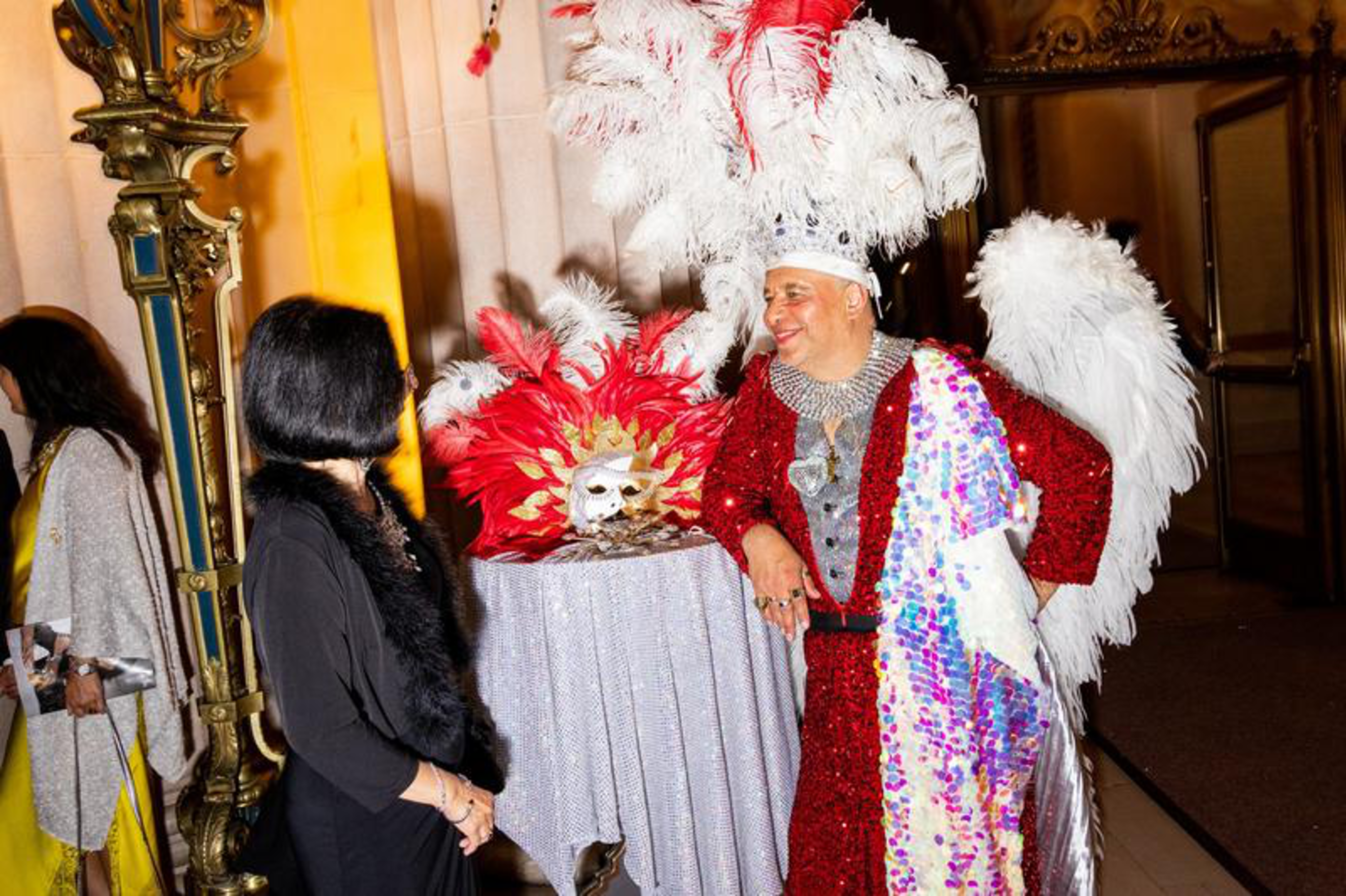 A man in a red sequined outfit with white feathered wings and a feathered headdress talks to a woman in black near a table with a feathered mask.
