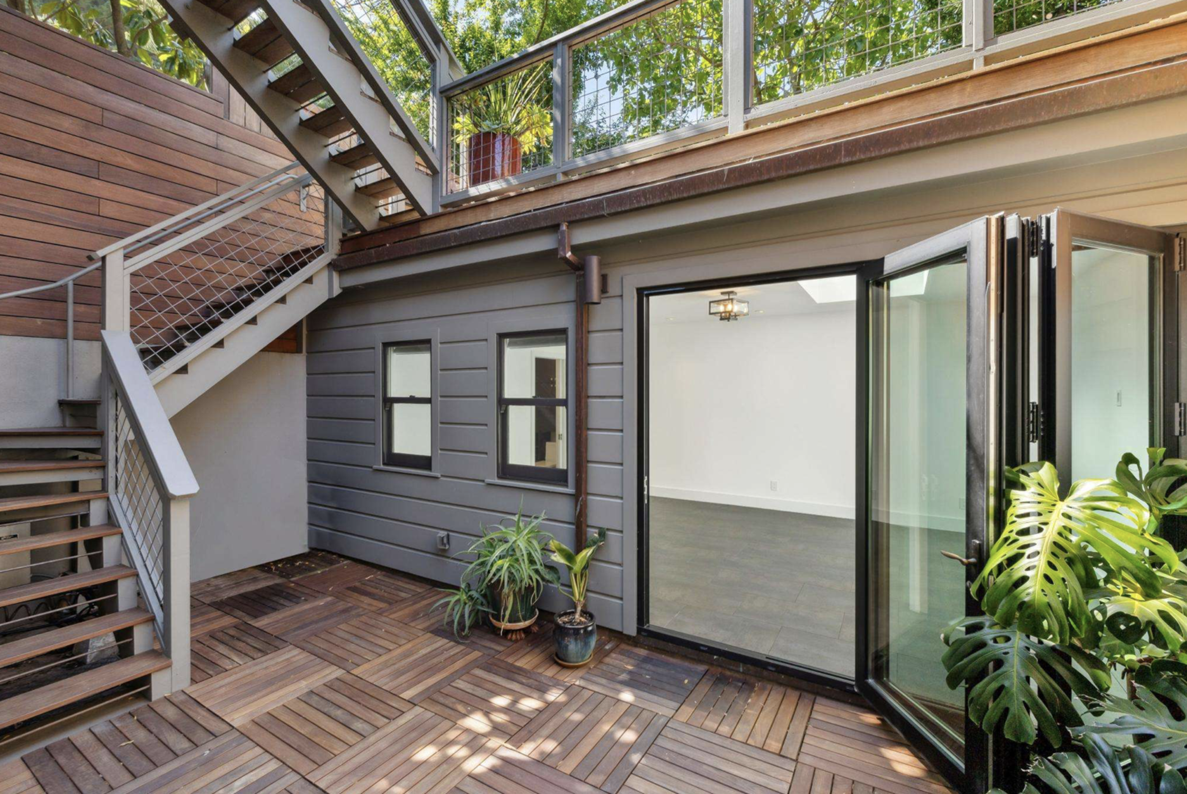 A wooden deck with potted plants leads to glass folding doors opening into a bright, empty room, with stairs ascending to an upper outdoor area.
