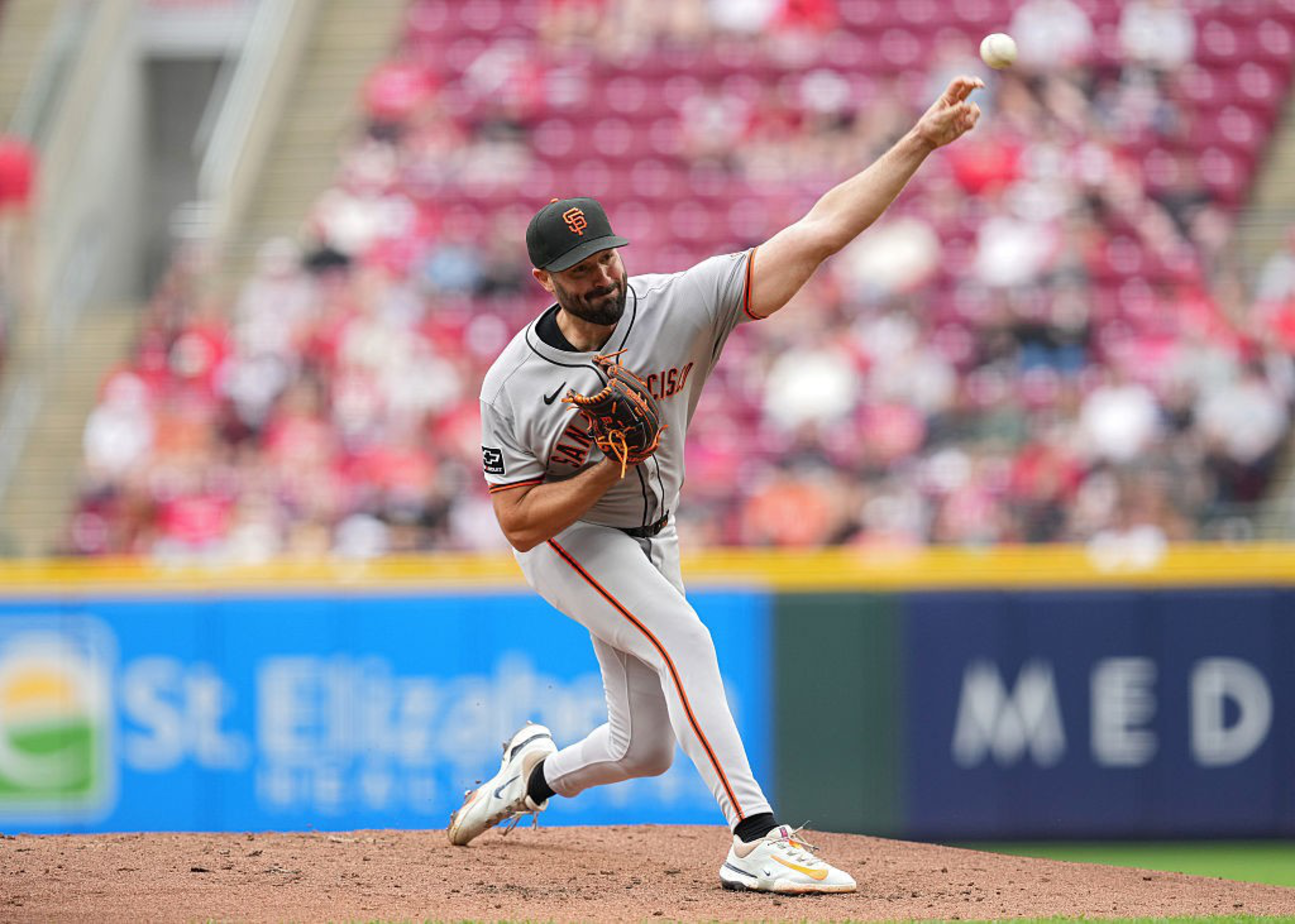 A baseball player in a gray uniform is pitching on a mound. The background shows a blurred crowd in red seats, and the player is mid-throw with the ball in the air.