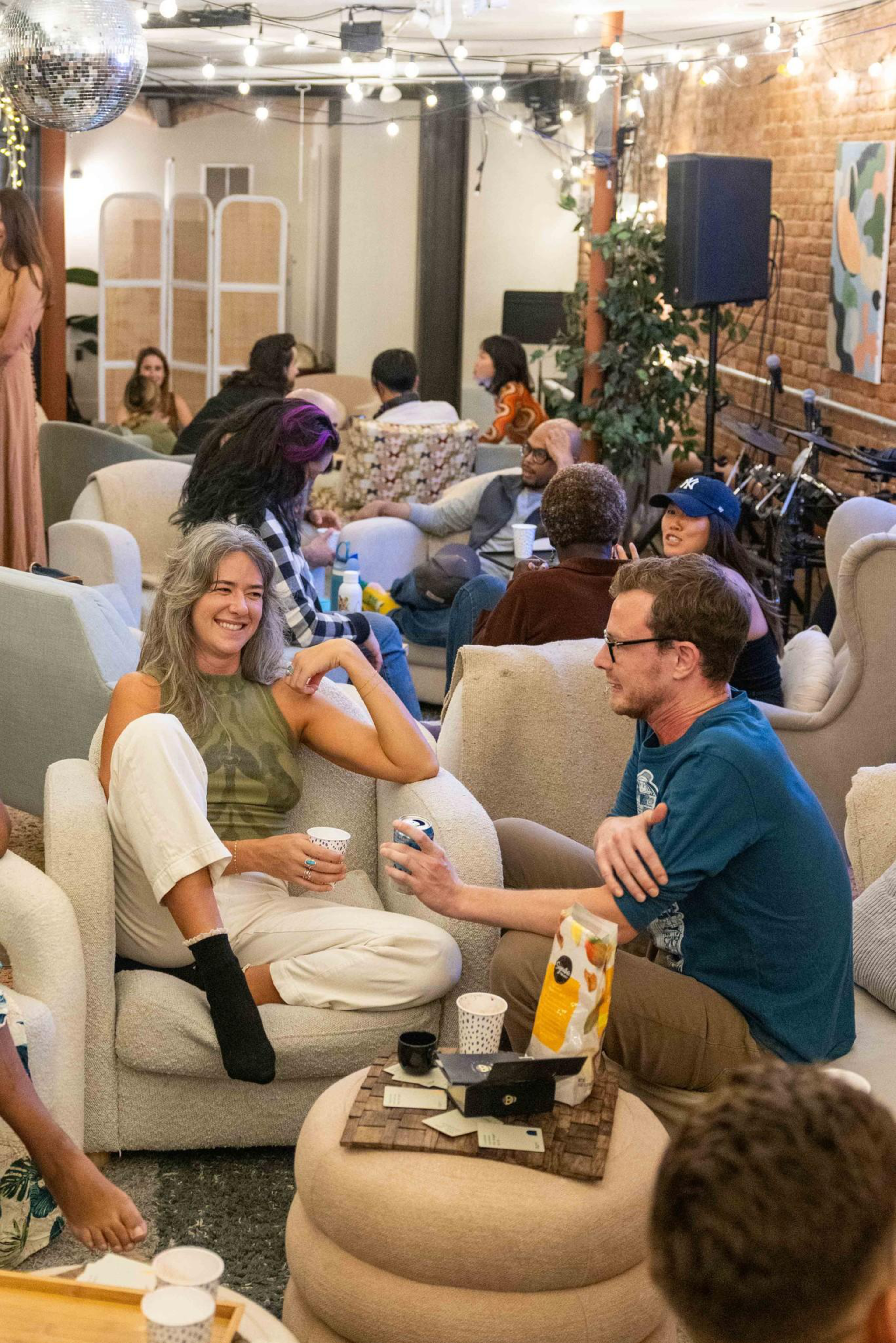 People are seated in cozy chairs, chatting and smiling in a warmly lit room decorated with string lights and a disco ball.