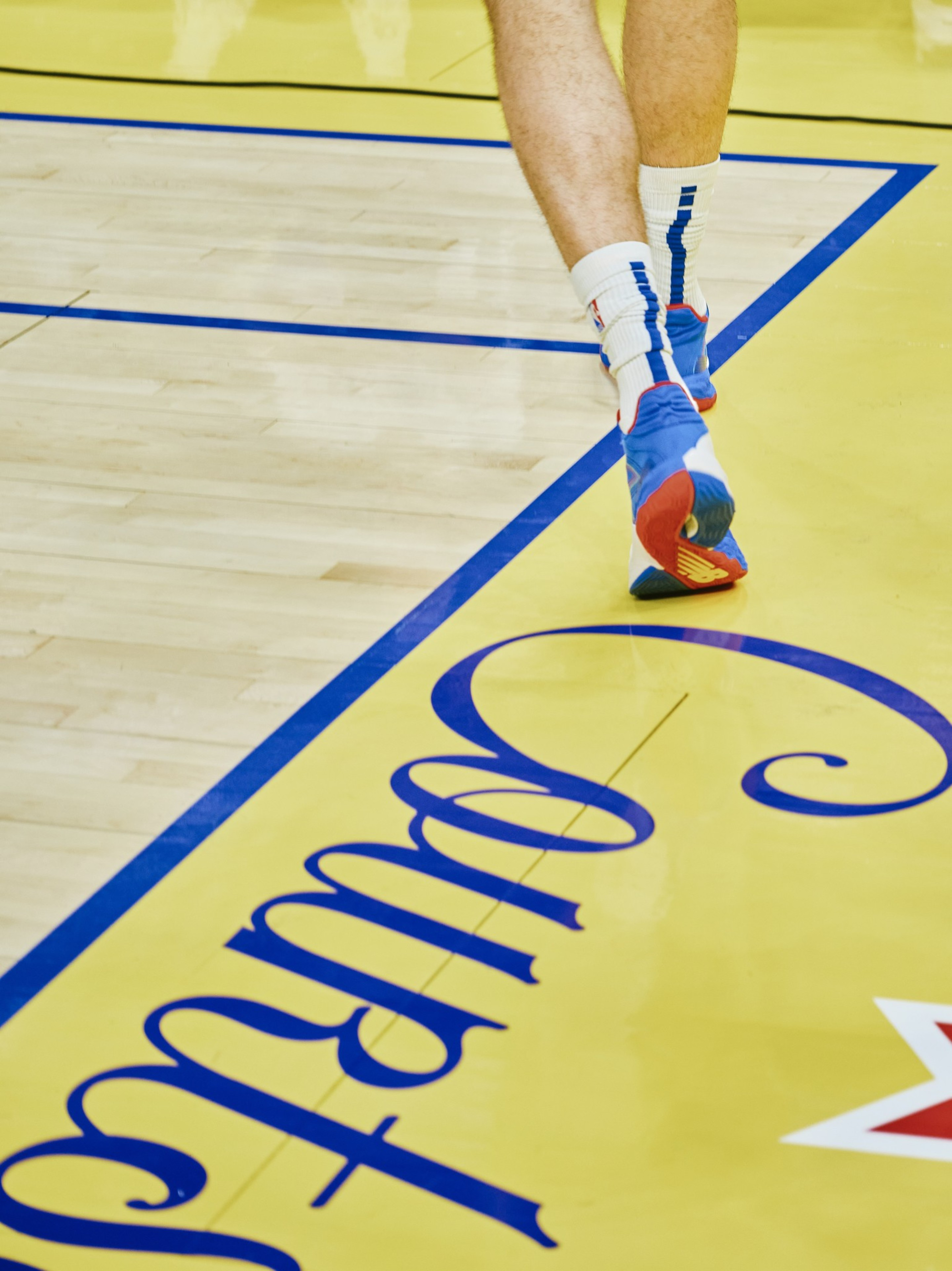 A person walks on a basketball court wearing blue and red sneakers with white socks. The court is yellow with blue lines and partial text visible.