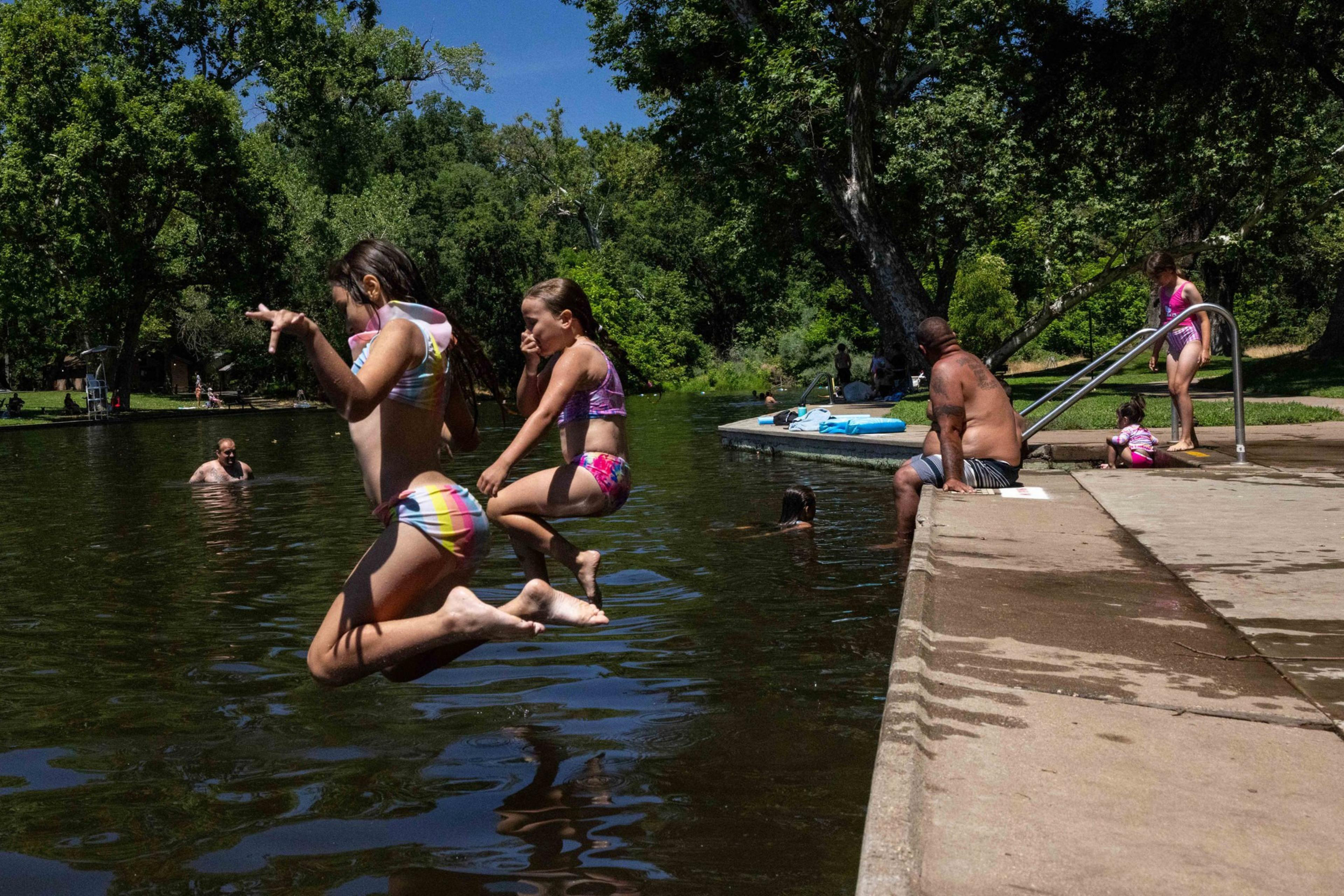 Two girls jump into a lake; nearby, a man and two kids sit on a concrete edge. Trees and other people enjoying the water are visible in the background.