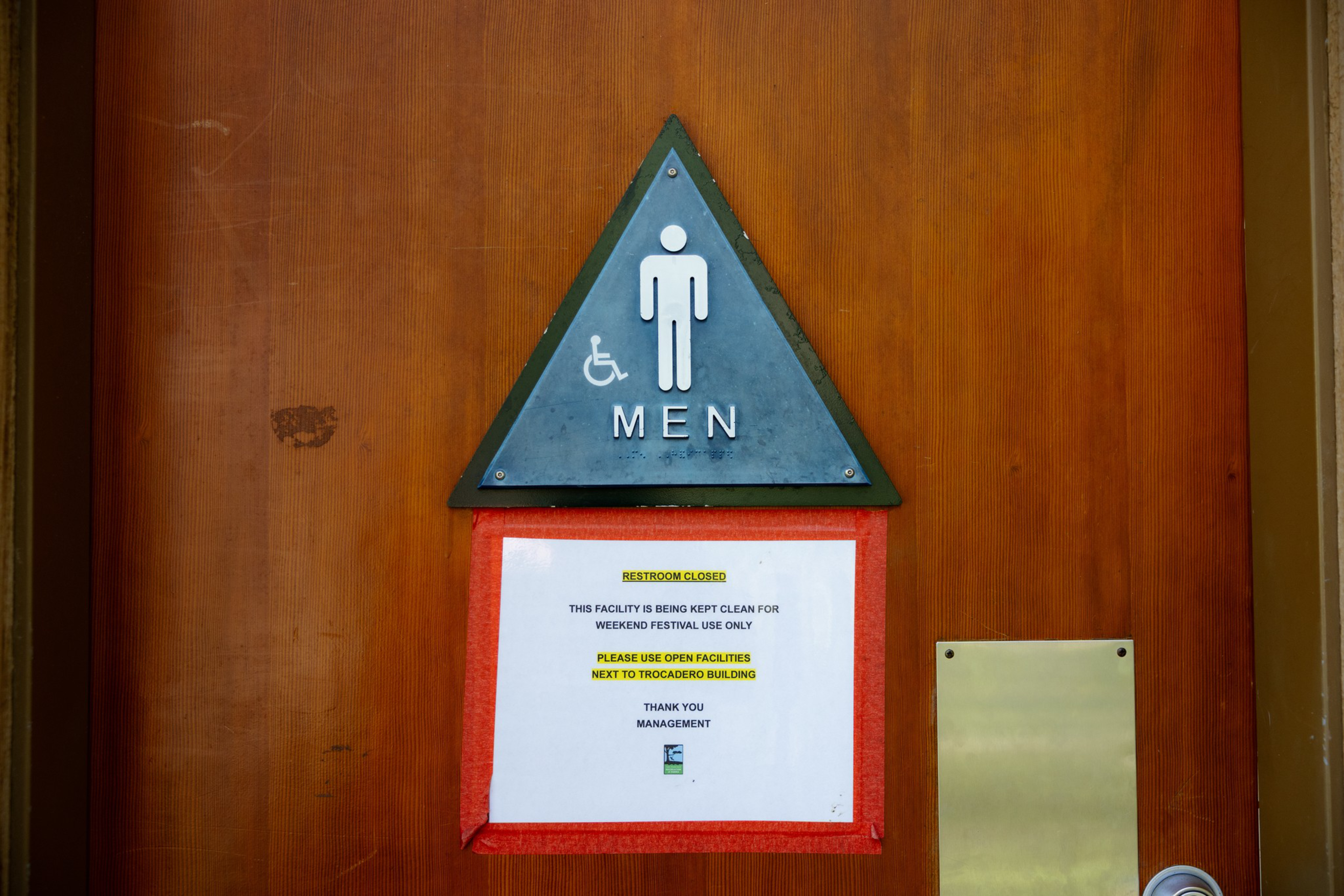 A wooden door with a triangular blue men’s restroom sign including a wheelchair symbol has a white notice below stating the restroom is closed for a weekend festival.